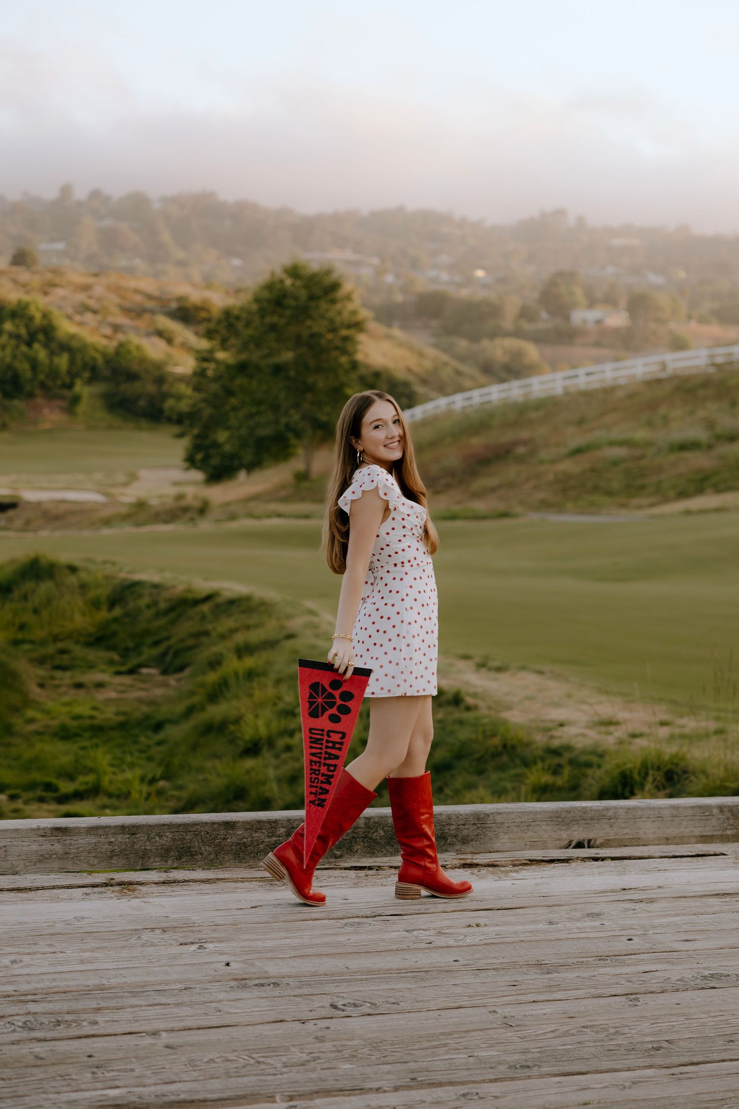 Young woman in polka dot dress and red boots holding a pennant, posing on wooden deck with scenic green hills.