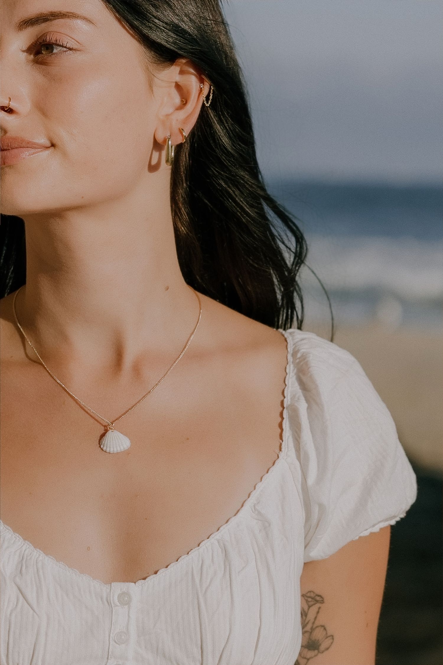 Woman wearing a seashell pendant necklace and gold hoop earrings with a white dress near the beach.