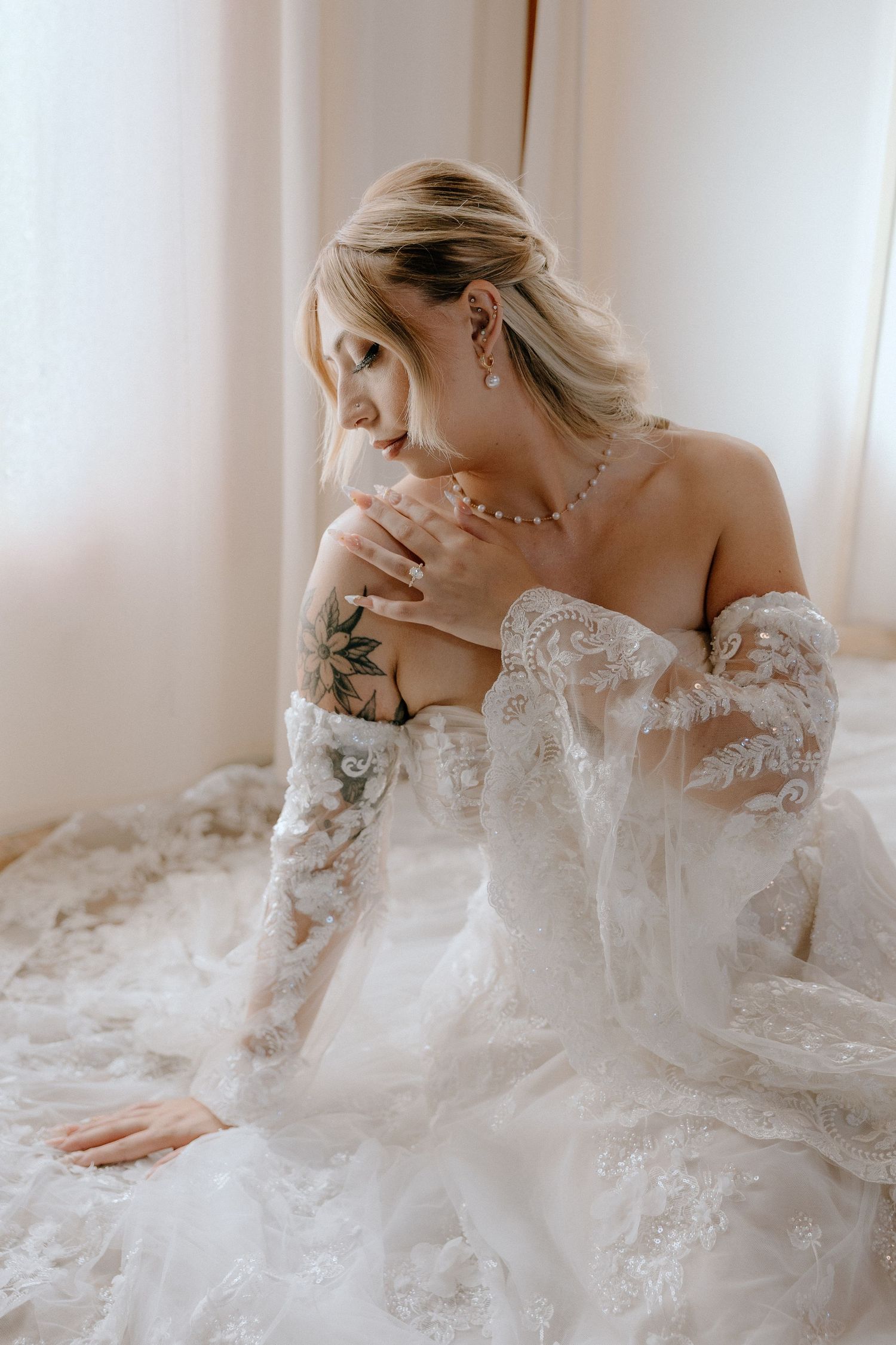 Bride in off-shoulder lace wedding gown with tattoo, pearl necklace, sitting gracefully in soft natural light.