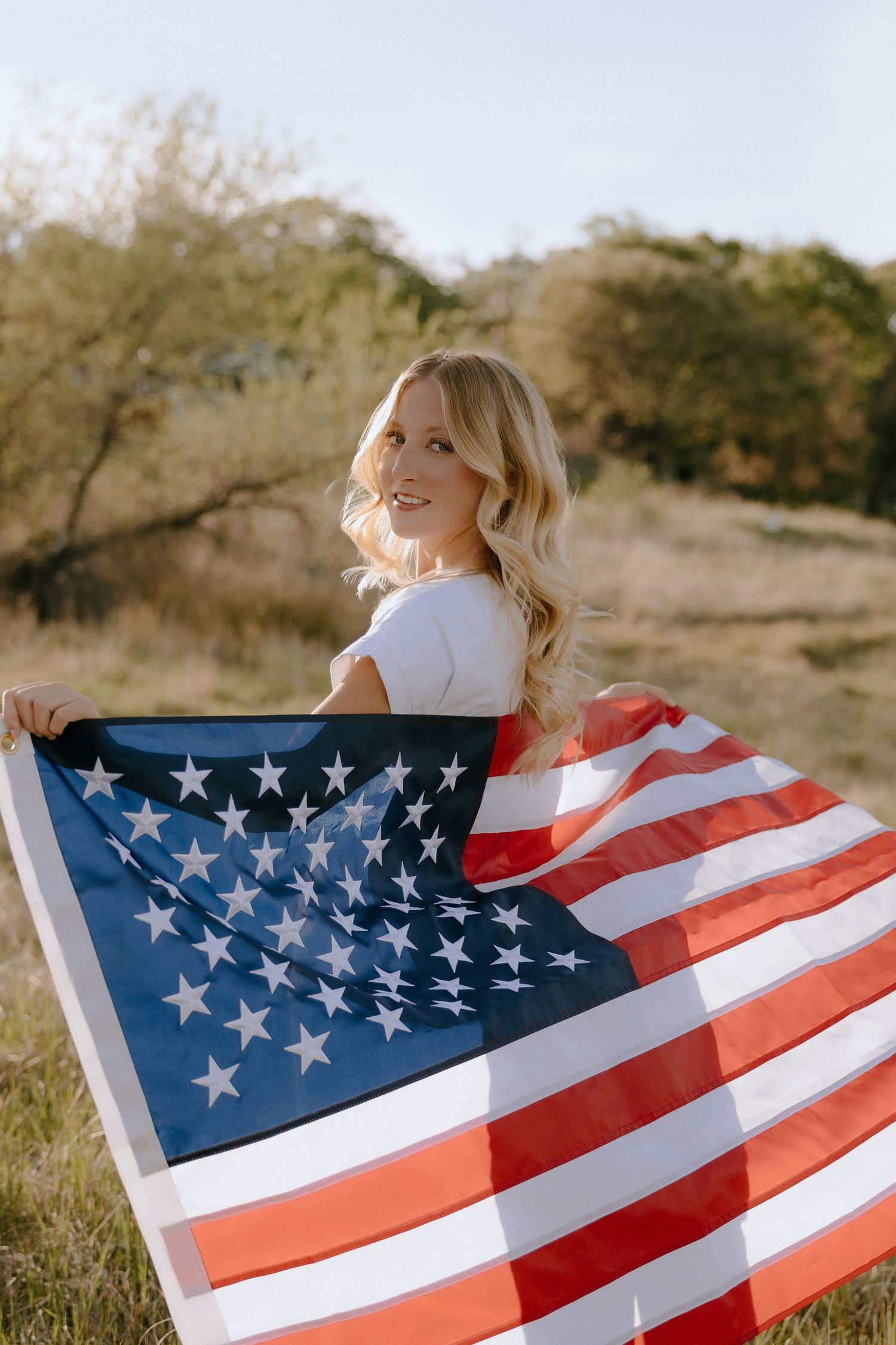 Smiling blonde woman holding a large American flag outdoors in a sunny field.