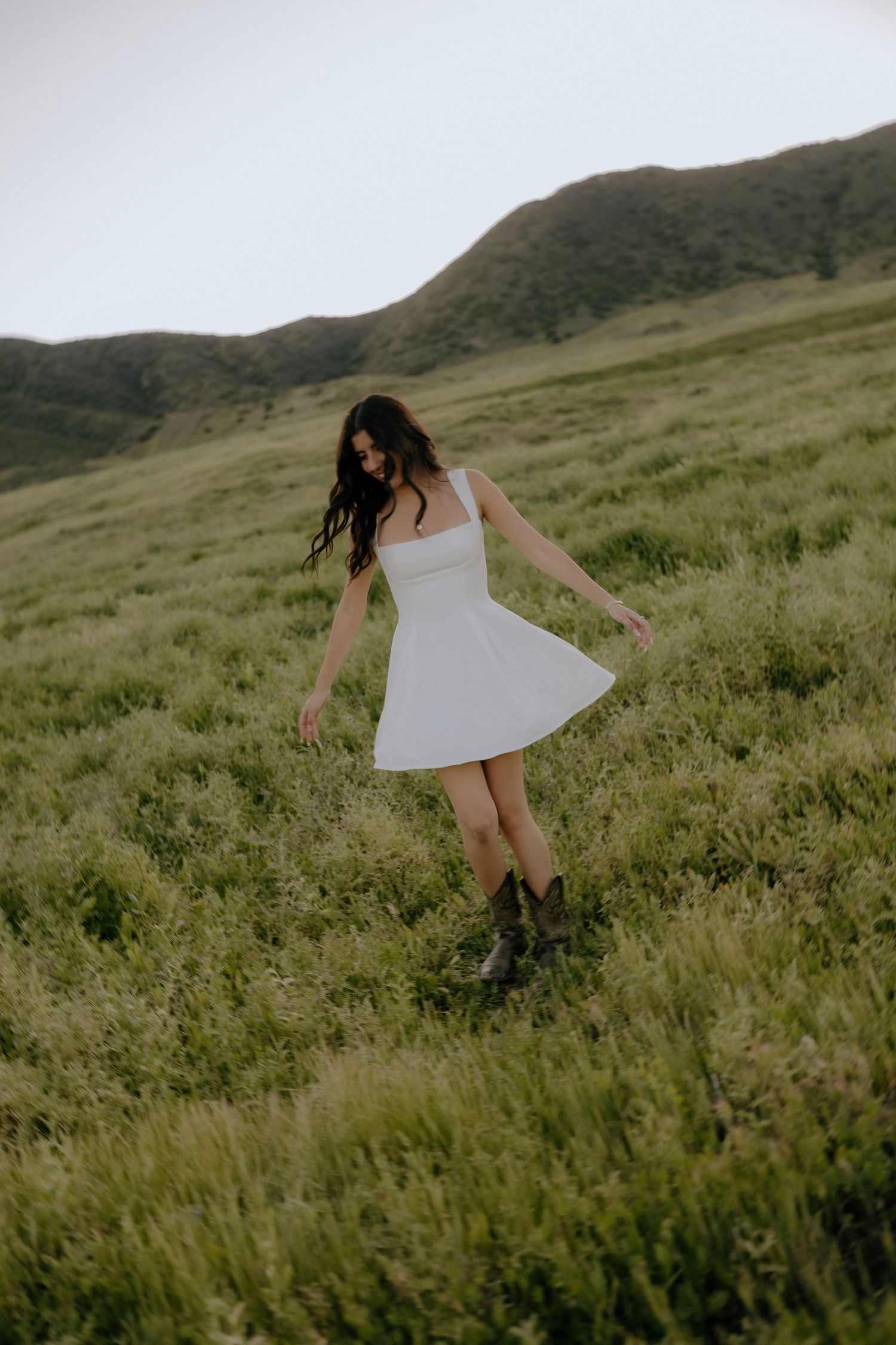 A woman in a white sundress and cowboy boots walks through a lush green hillside meadow.