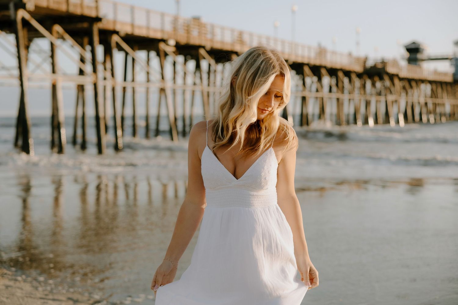 Blonde woman in white sundress walking on beach near wooden pier in golden hour light.