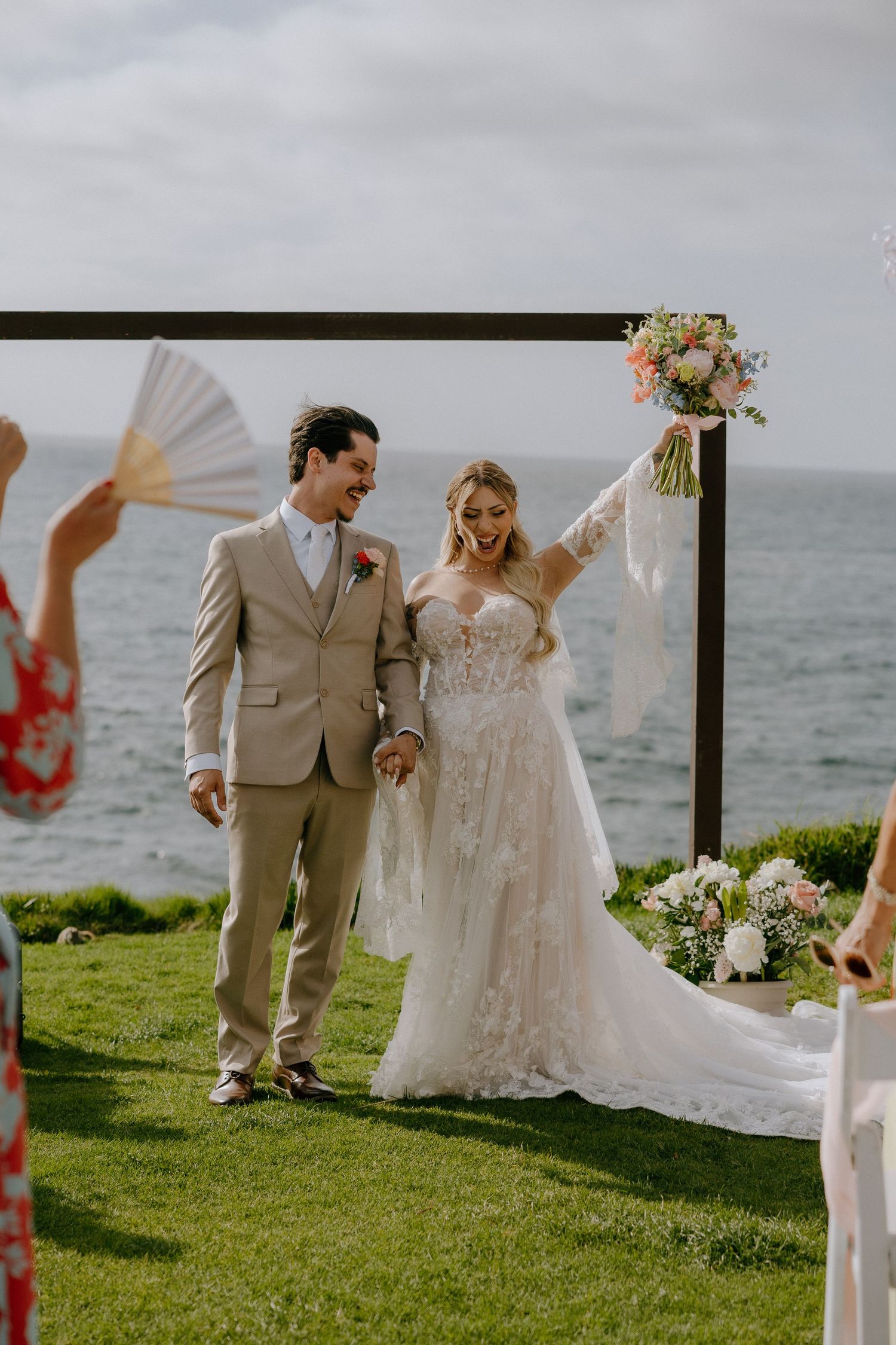 Joyful bride in lace gown raises bouquet beside groom in tan suit at outdoor oceanfront wedding ceremony.