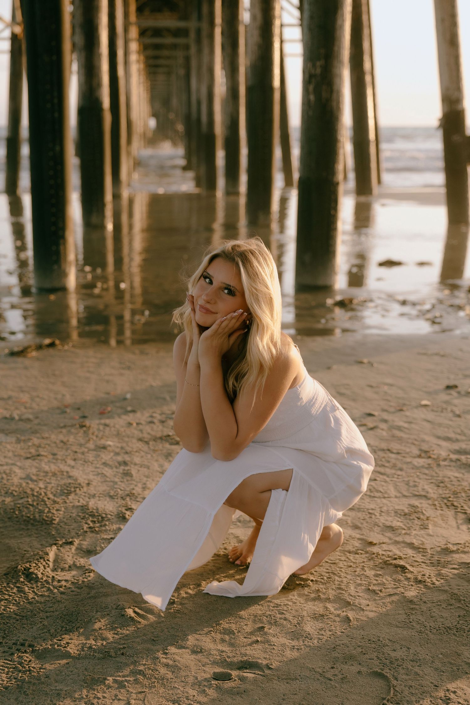 Blonde woman in white dress crouching on sandy beach under wooden pier at golden hour.