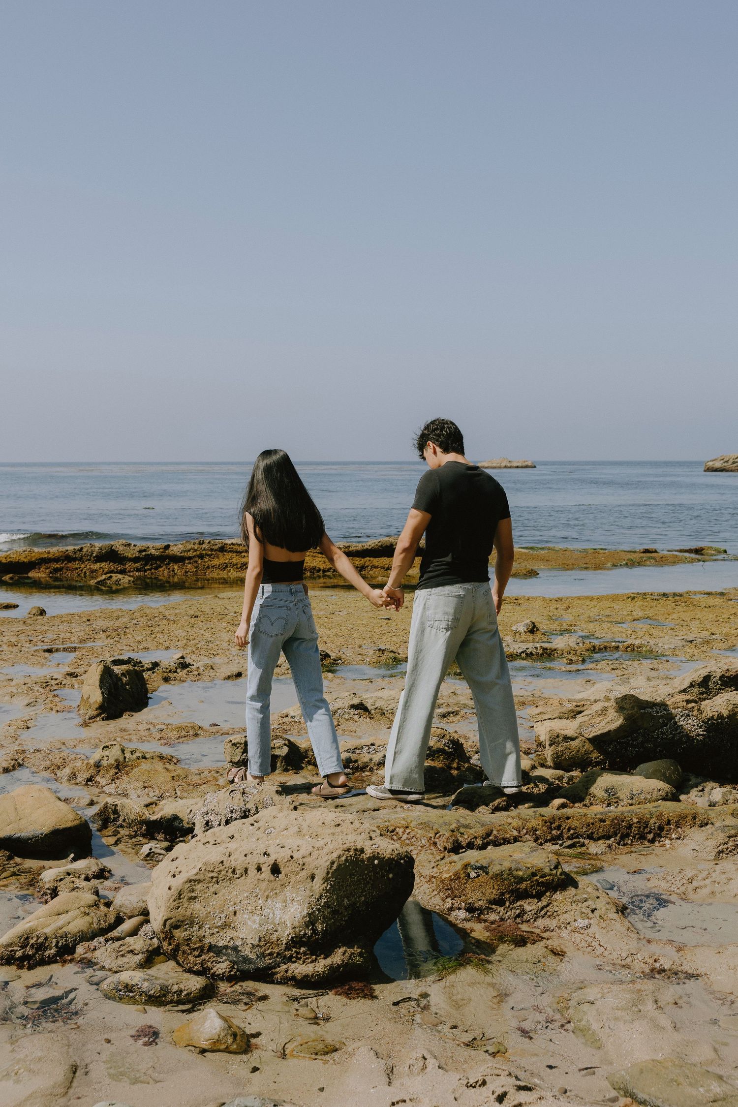 Couple holding hands walking on rocky beach shore with calm ocean and clear blue sky in background.