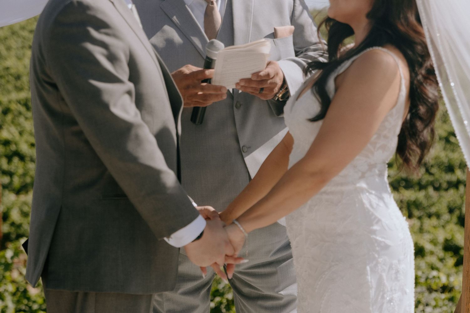 Bride and groom holding hands during outdoor wedding ceremony while officiant reads vows.