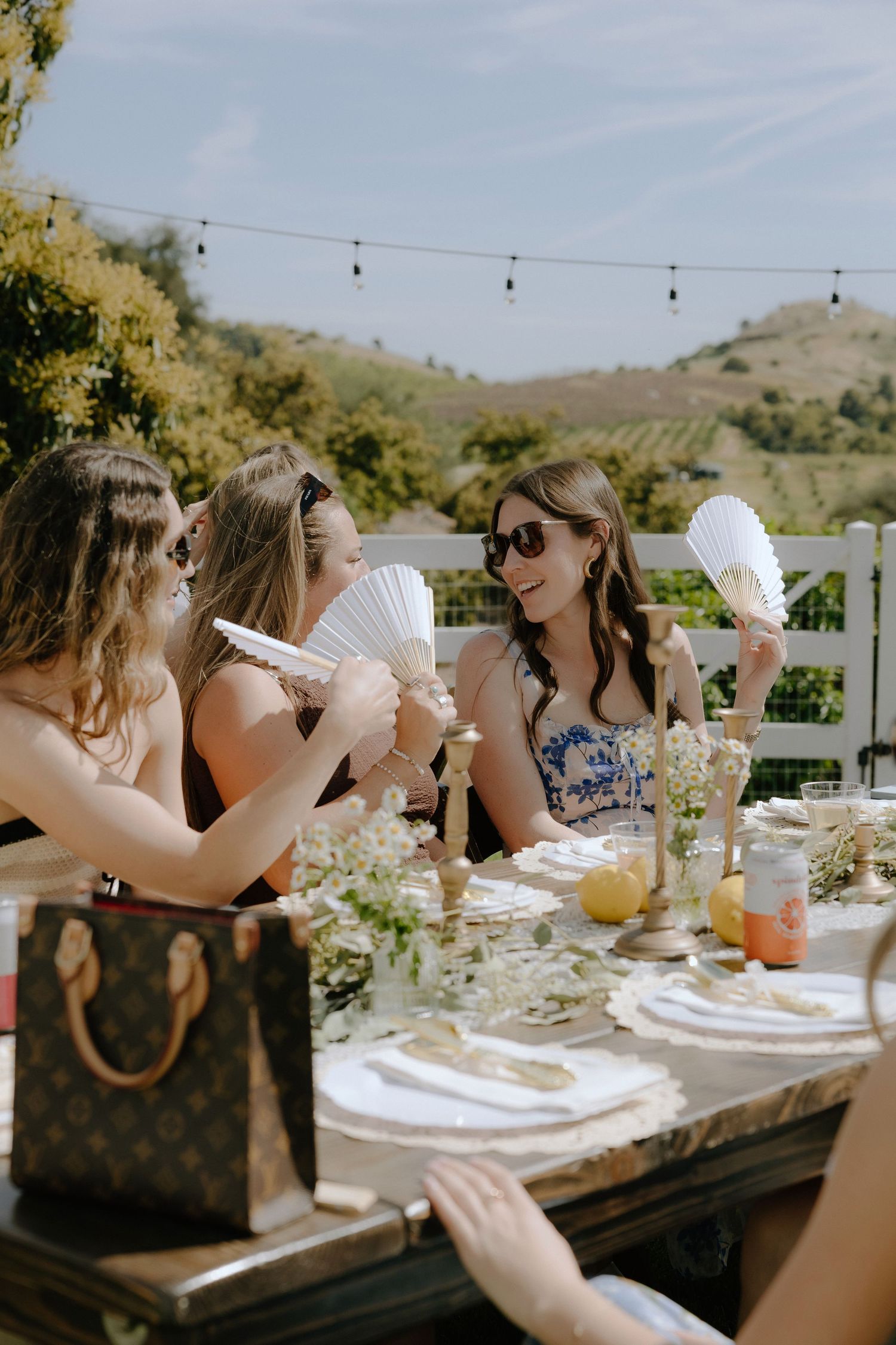 Women holding hand fans at an elegant outdoor garden party table with floral arrangements and rolling hills backdrop.