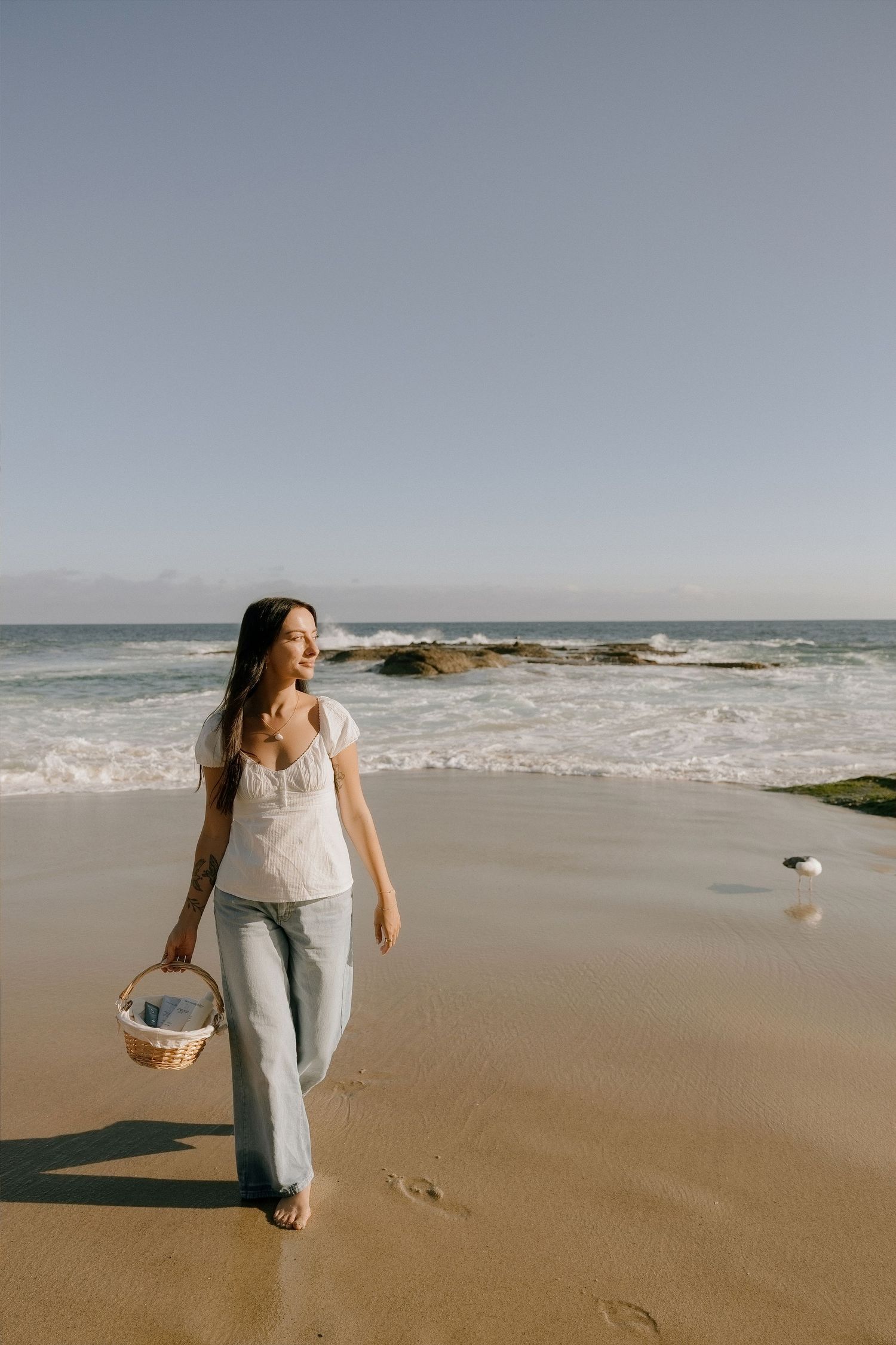 Woman walking barefoot on sandy beach carrying wicker basket, wearing white top and jeans with ocean waves behind her.