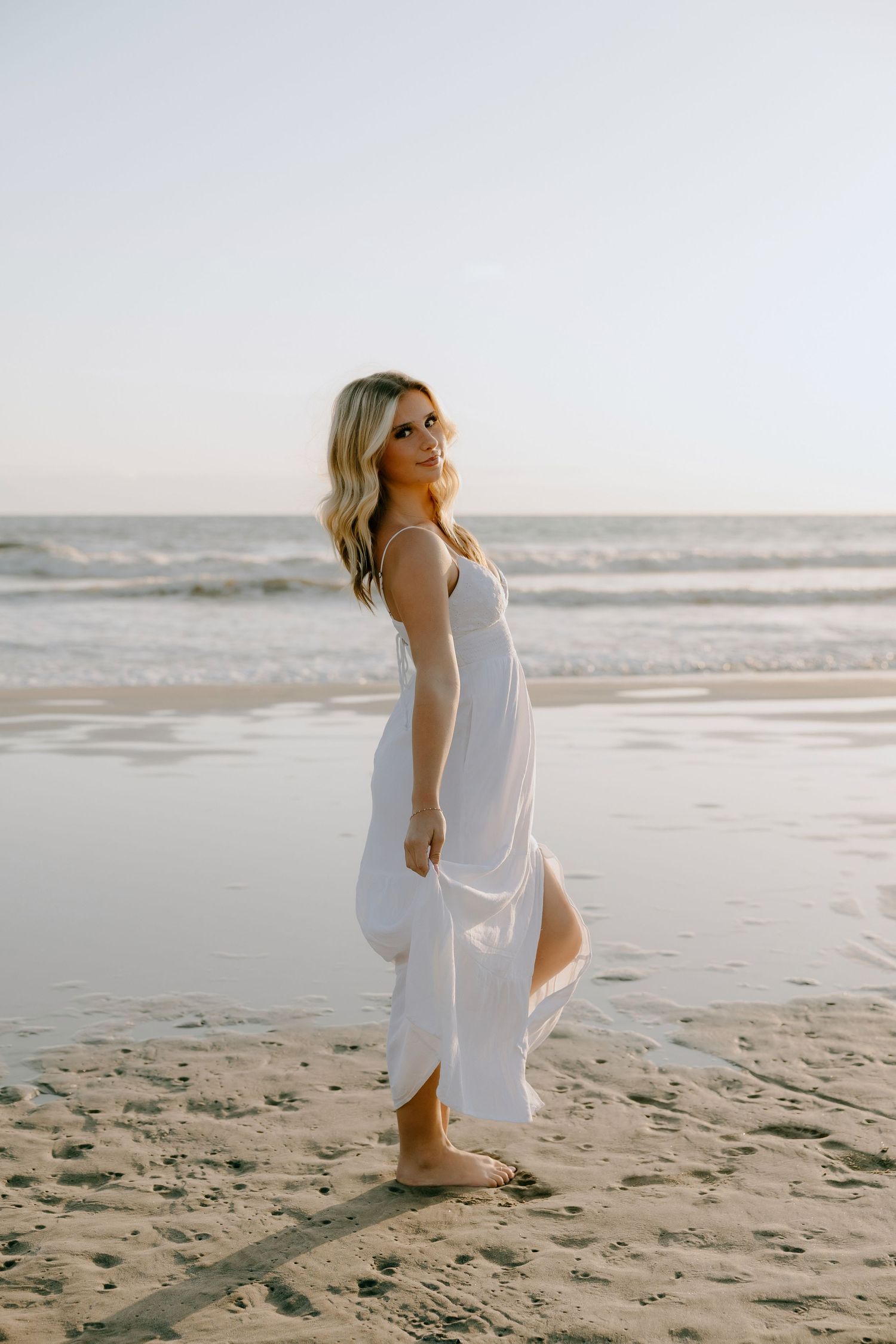 Blonde woman in white maxi dress walking barefoot on sandy beach at sunset with ocean waves behind her.
