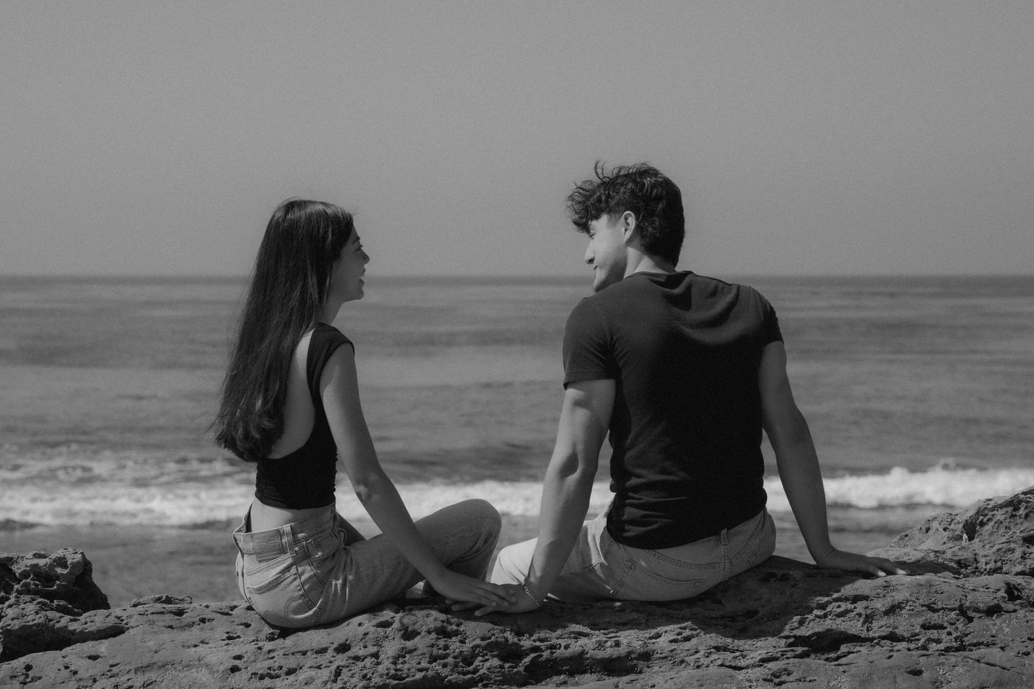 Black and white photo of a young couple sitting on rocks by the ocean, facing each other in conversation.