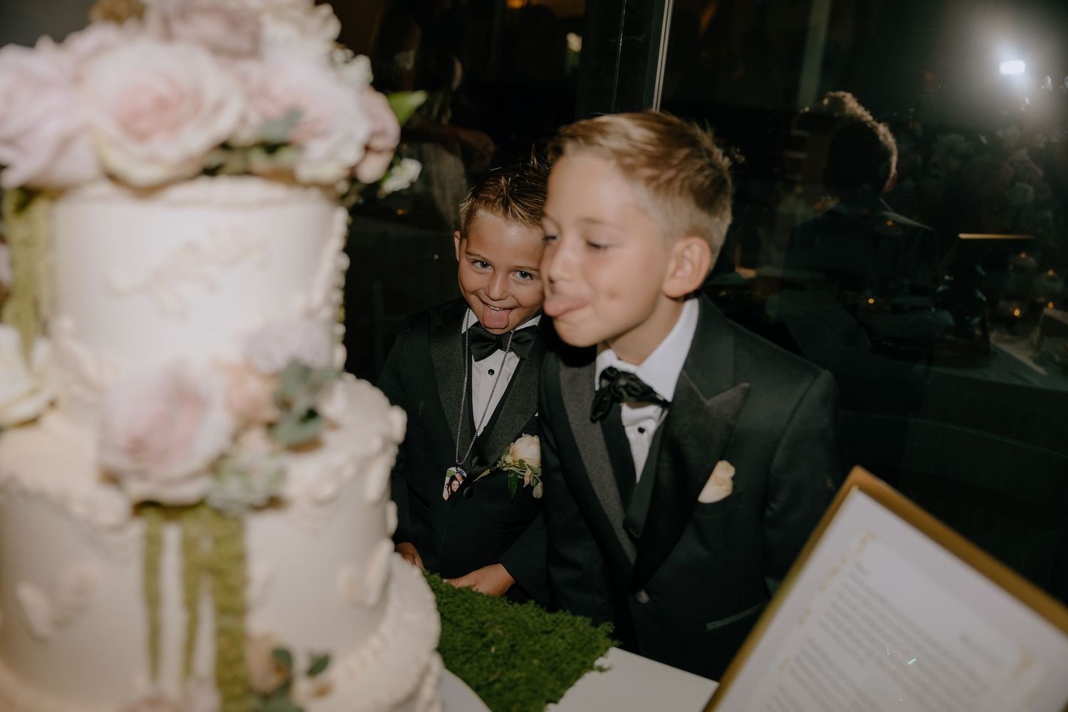 Two boys in black tuxedos admire a elegant white wedding cake adorned with pink roses.