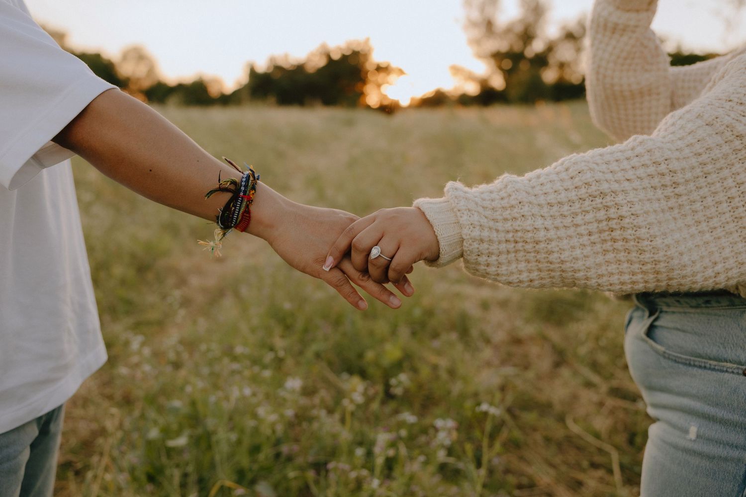 Two people holding hands in a golden field at sunset, one wearing a bracelet and a white top.