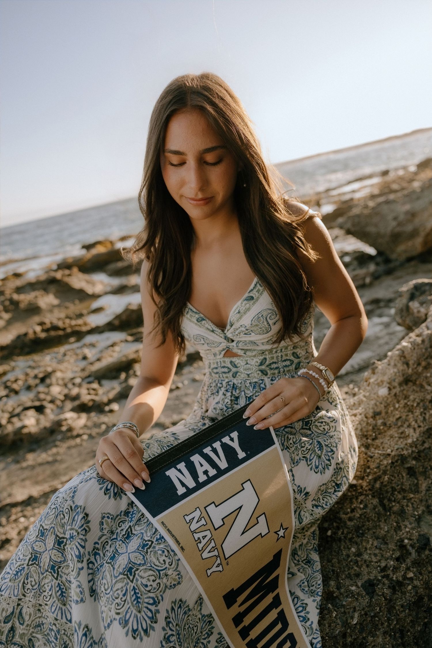 Young woman in floral dress holding Navy pennant on rocky shore.