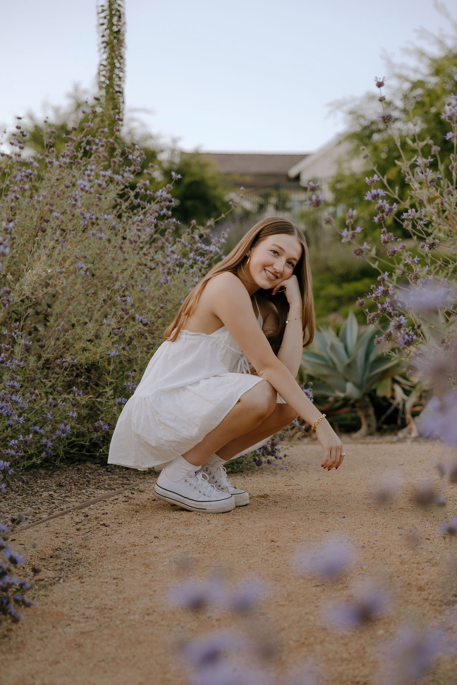 Young woman in white dress and sneakers crouching among purple wildflowers in a garden, smiling warmly.