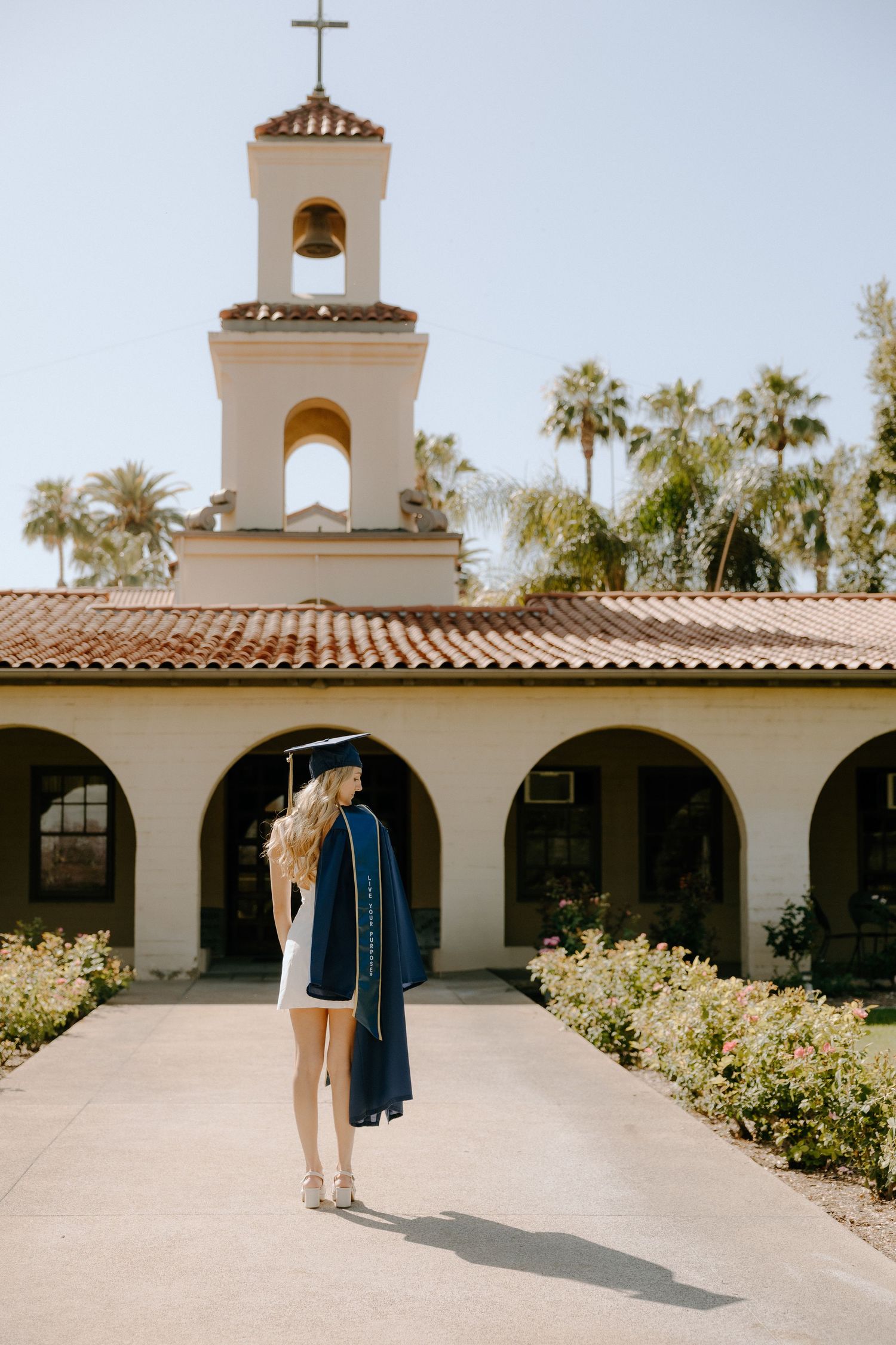 Graduate in cap and gown stands before a white Spanish-style chapel with bell tower and red tile roof on a sunny day.