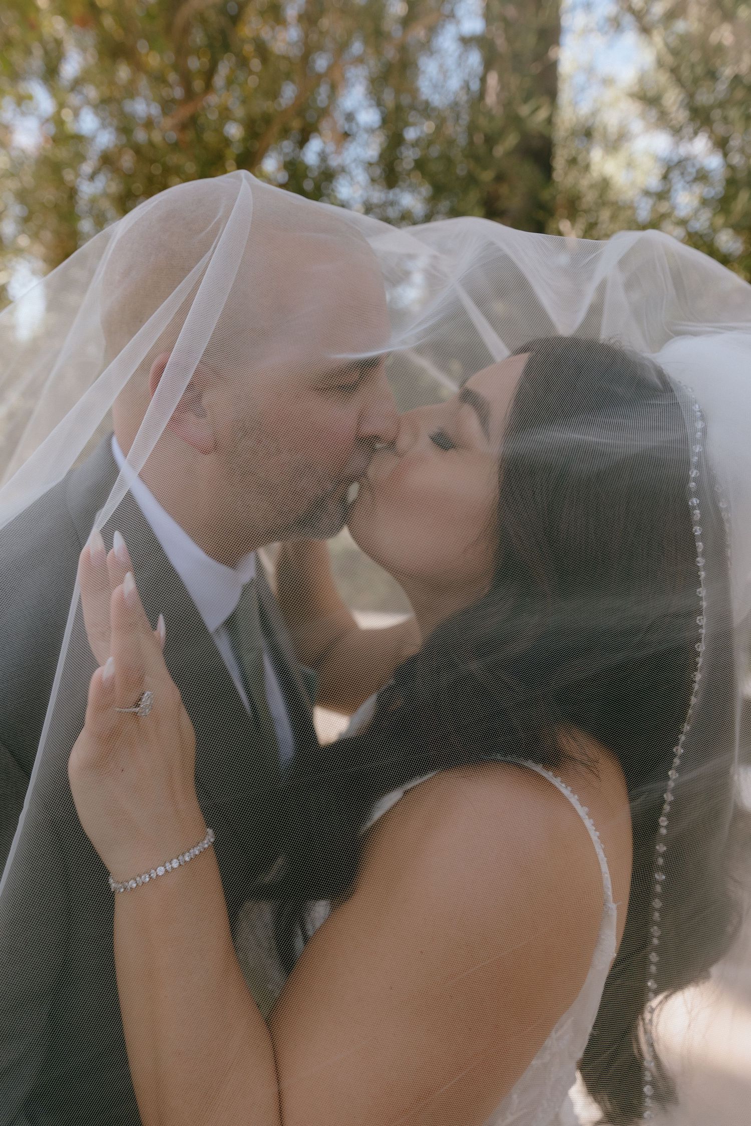 A bride and groom share a romantic kiss under a flowing wedding veil outdoors, surrounded by lush green trees.