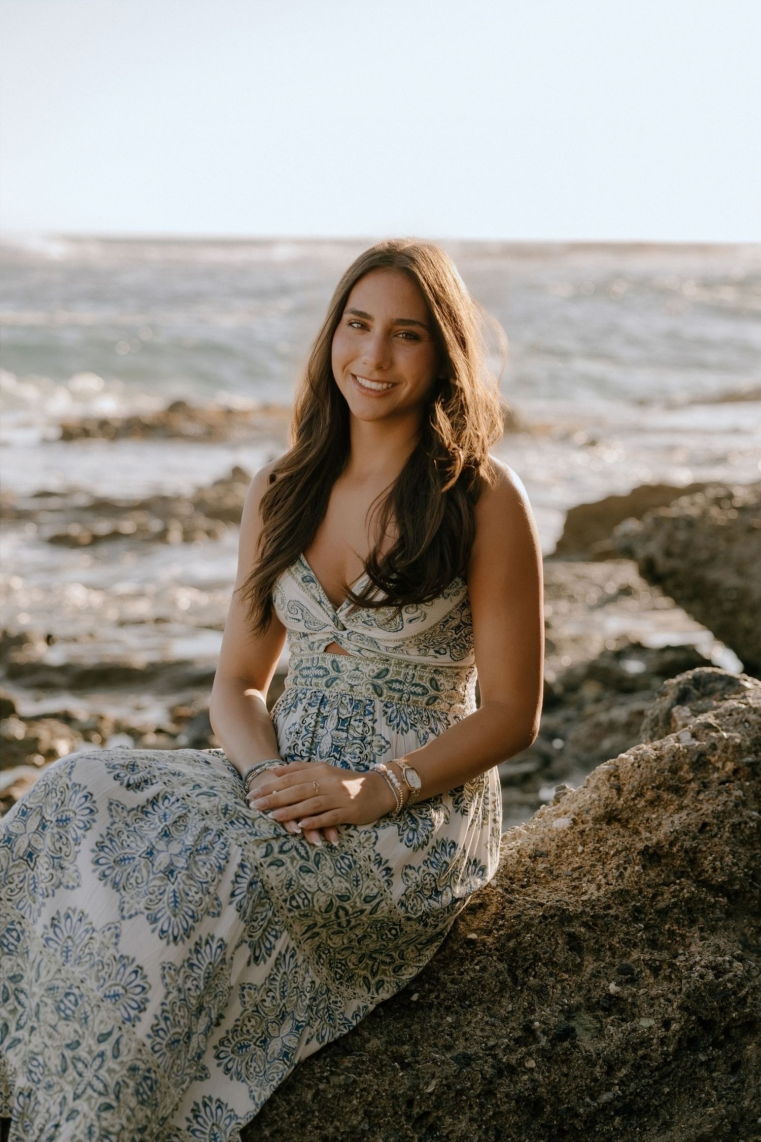 A smiling woman in a blue floral maxi dress sits on rocks by the ocean during golden hour.