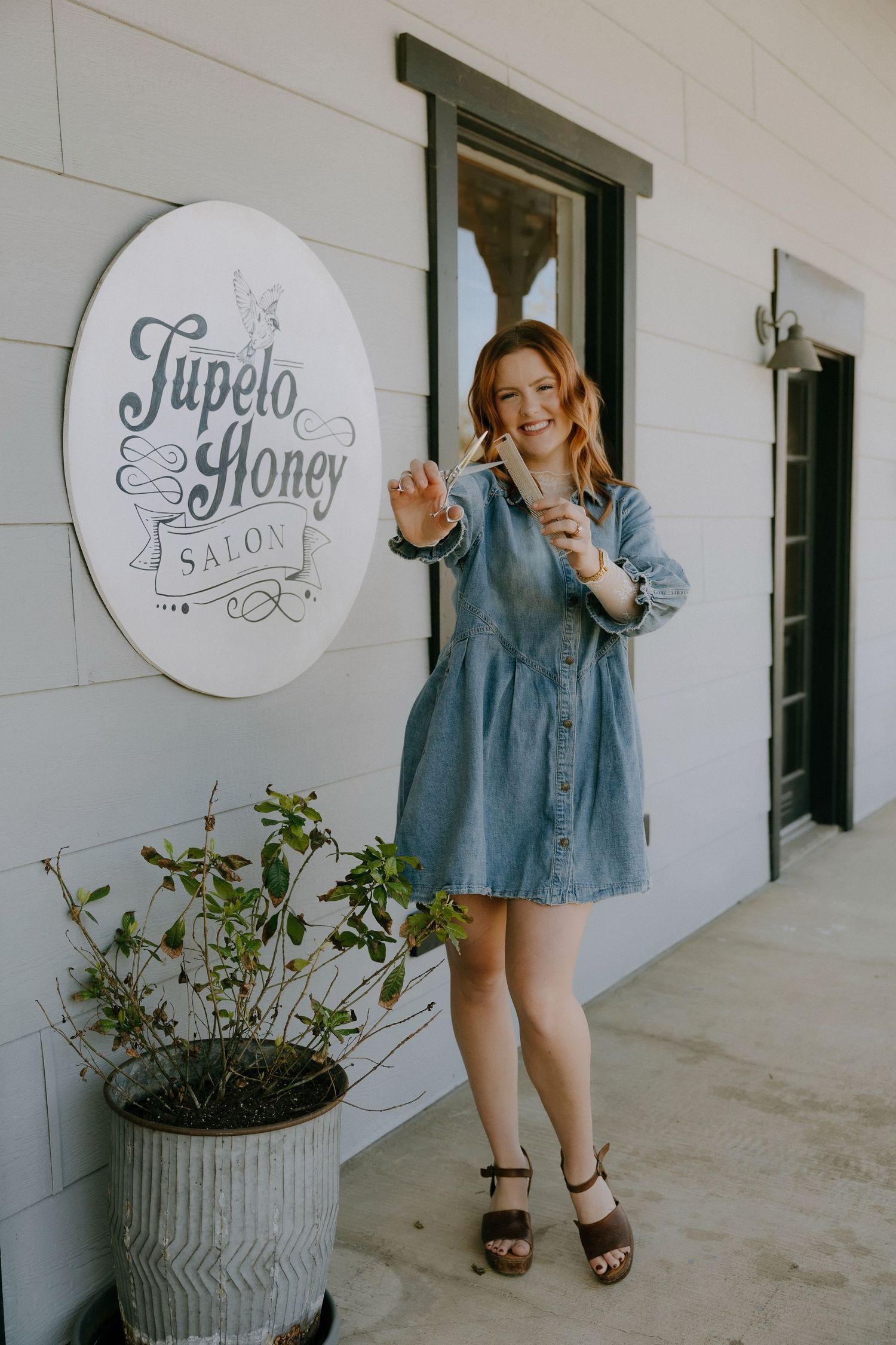 Woman in denim dress posing outside Tupelo Honey Salon holding scissors, wearing wooden heels.