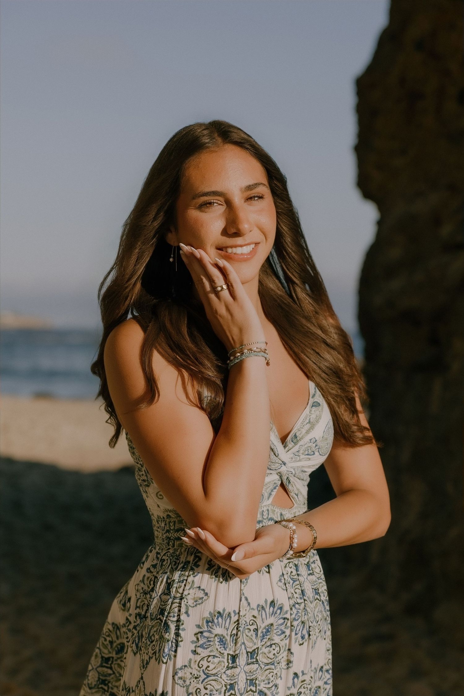 Smiling brunette woman in floral sundress poses at golden hour on a scenic beach.