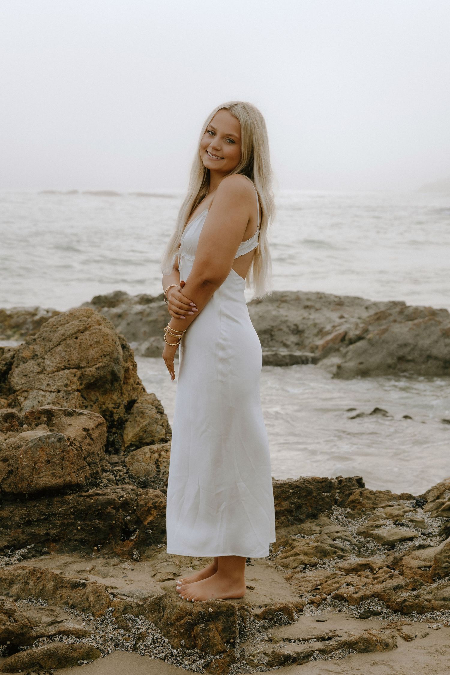 Smiling blonde woman in white midi dress standing barefoot on rocky beach with ocean waves in background.