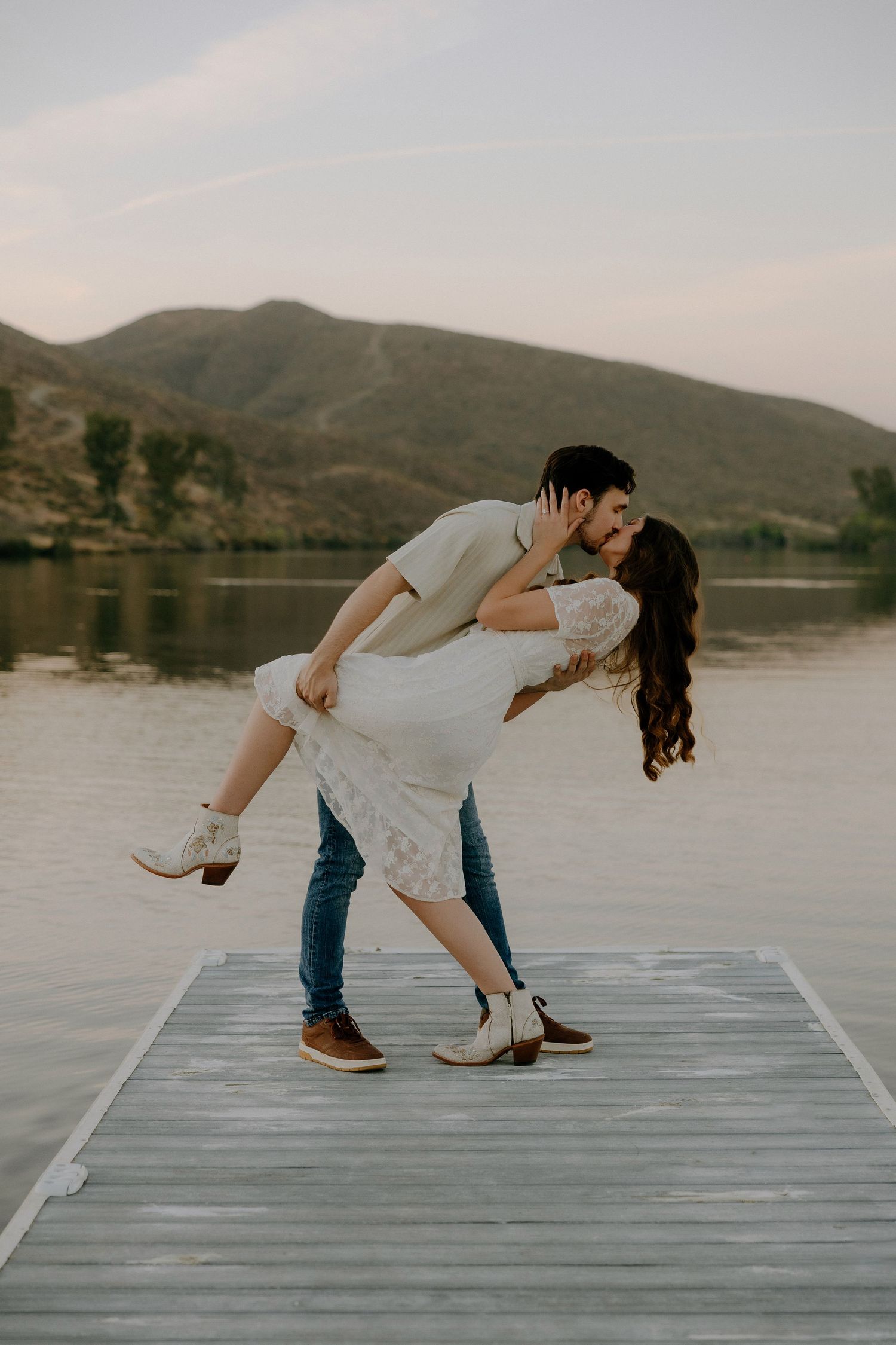 Couple sharing a romantic dip kiss on a wooden dock by a serene lake with mountain backdrop at golden hour.