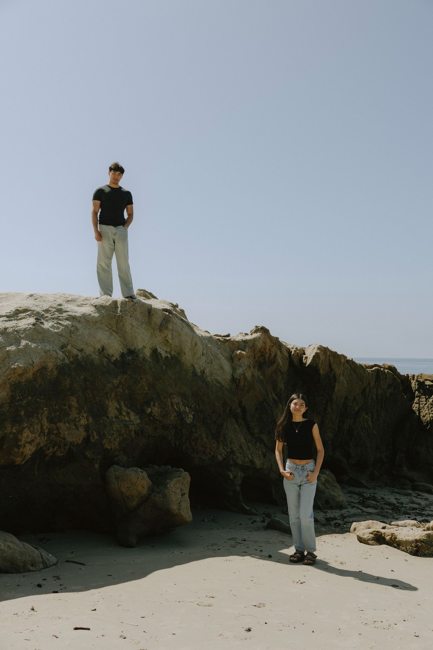 A man stands atop a rocky cliff while a woman looks up at him on a sandy beach by the ocean.