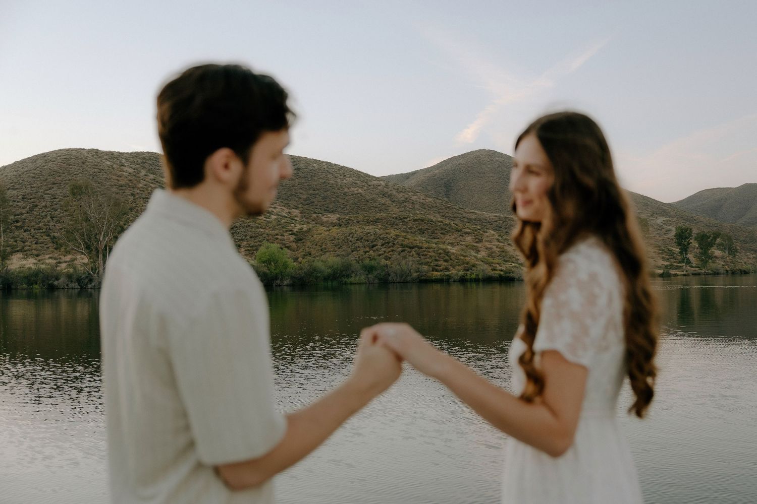 A couple holds hands by a serene lake with rolling hills at dusk, sharing a romantic outdoor moment.