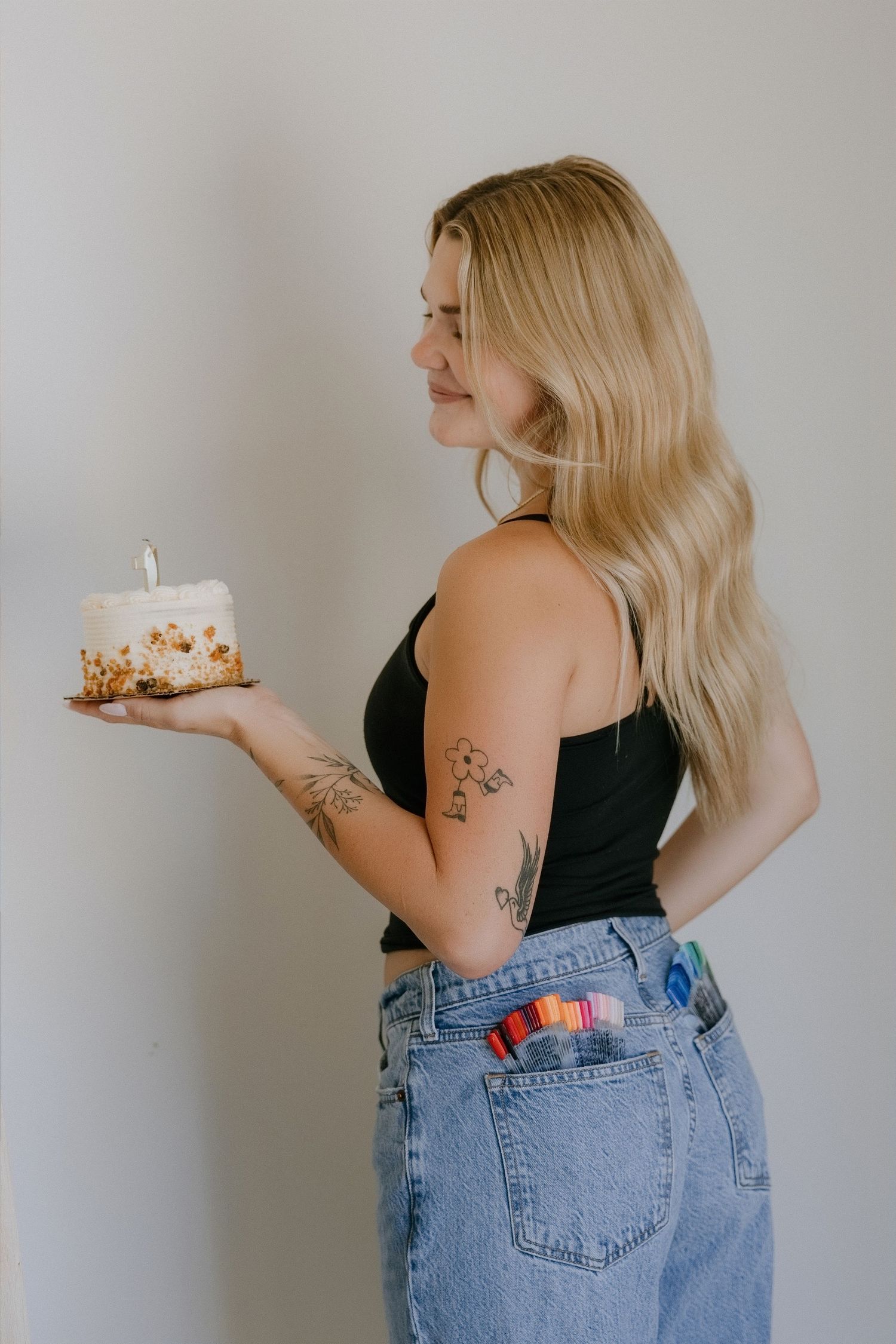 Blonde woman with tattoos holding a small birthday cake with a candle, wearing black top and jeans.