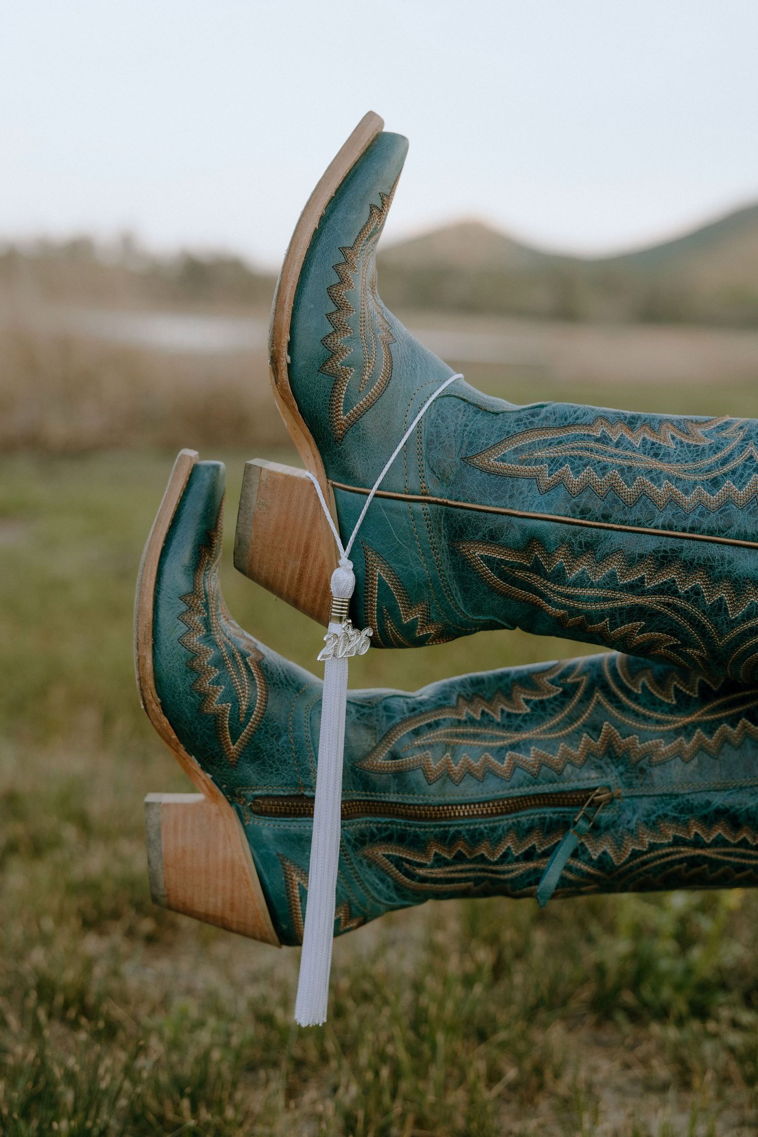 Teal cowboy boots with tan stitching displayed outdoors, adorned with a white tassel charm, set against a scenic landscape.