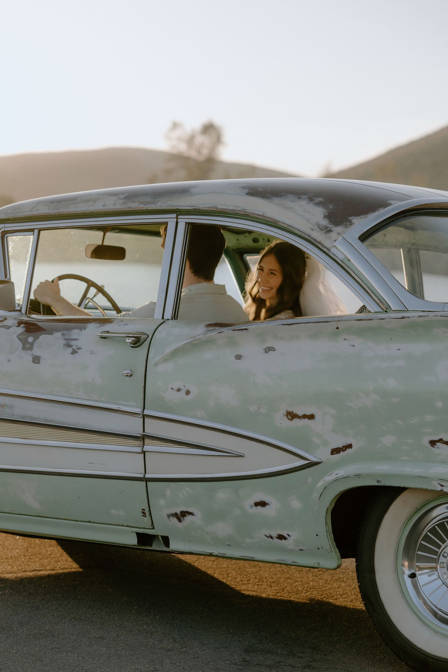 A couple sits in a vintage mint green classic car at golden hour with rolling hills in the background.