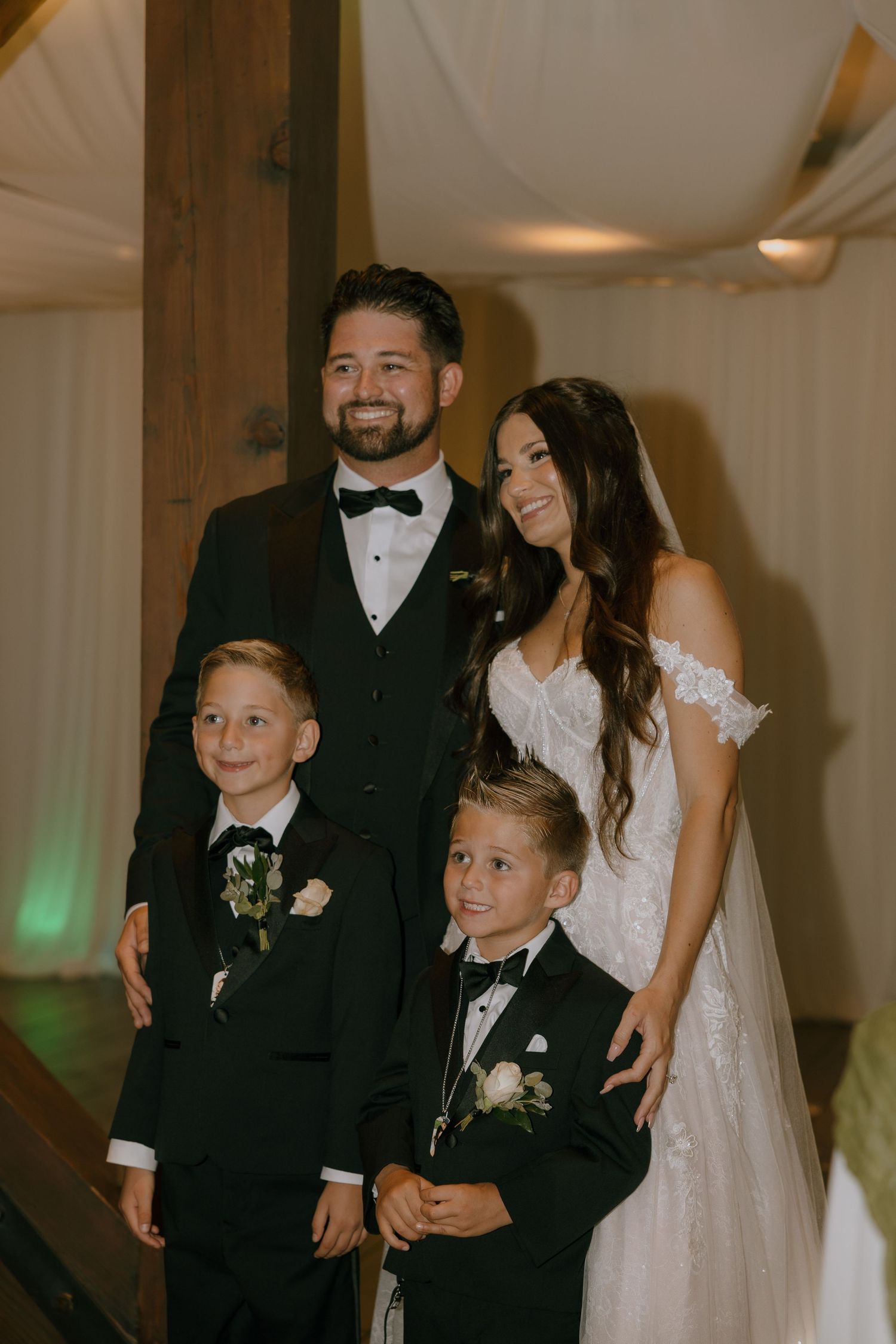 A smiling bride and groom pose with two young boys in tuxedos at an elegant wedding reception.