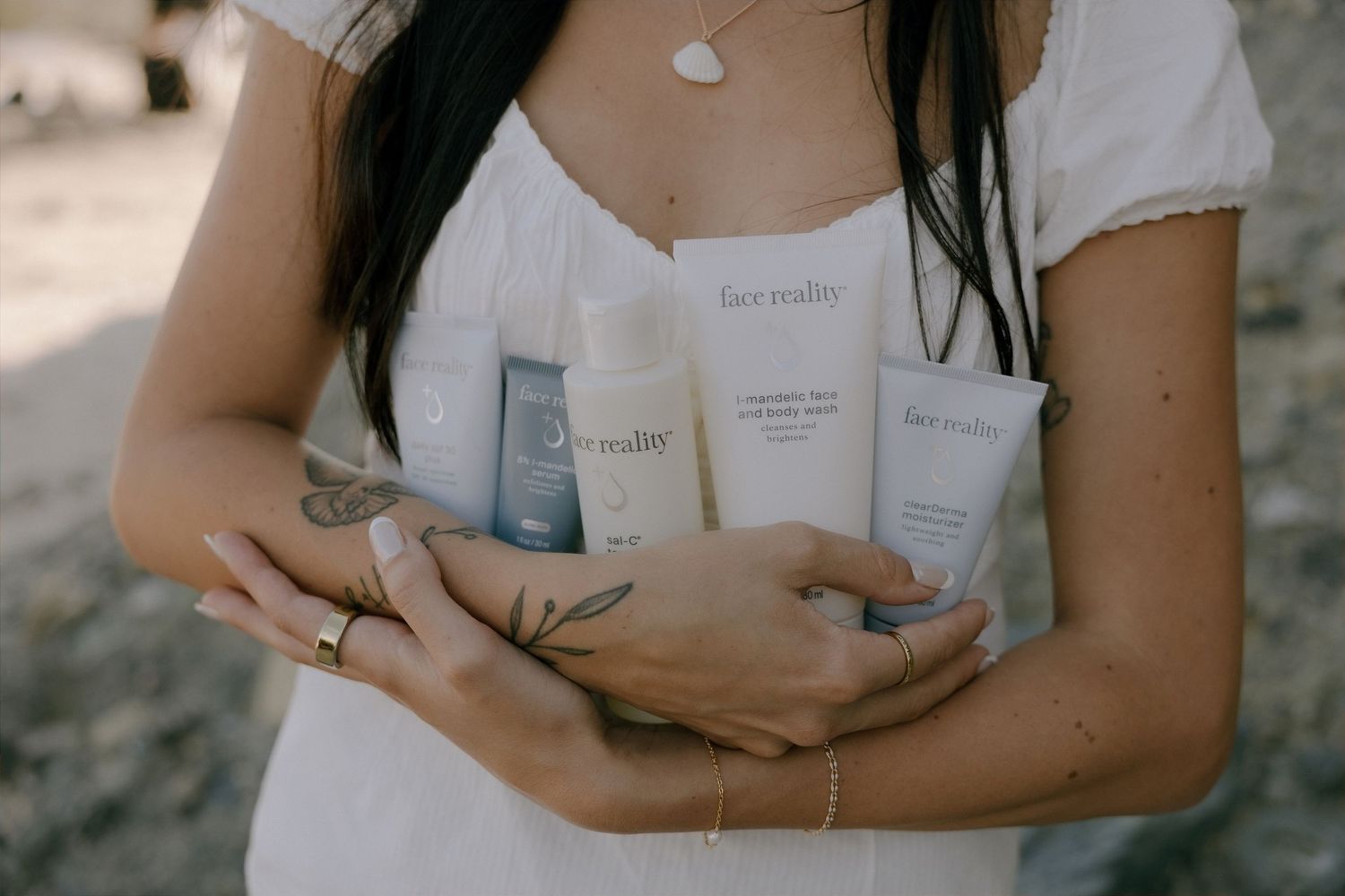 Woman in white dress holding Face Reality skincare products including cleanser and moisturizer on a beach.
