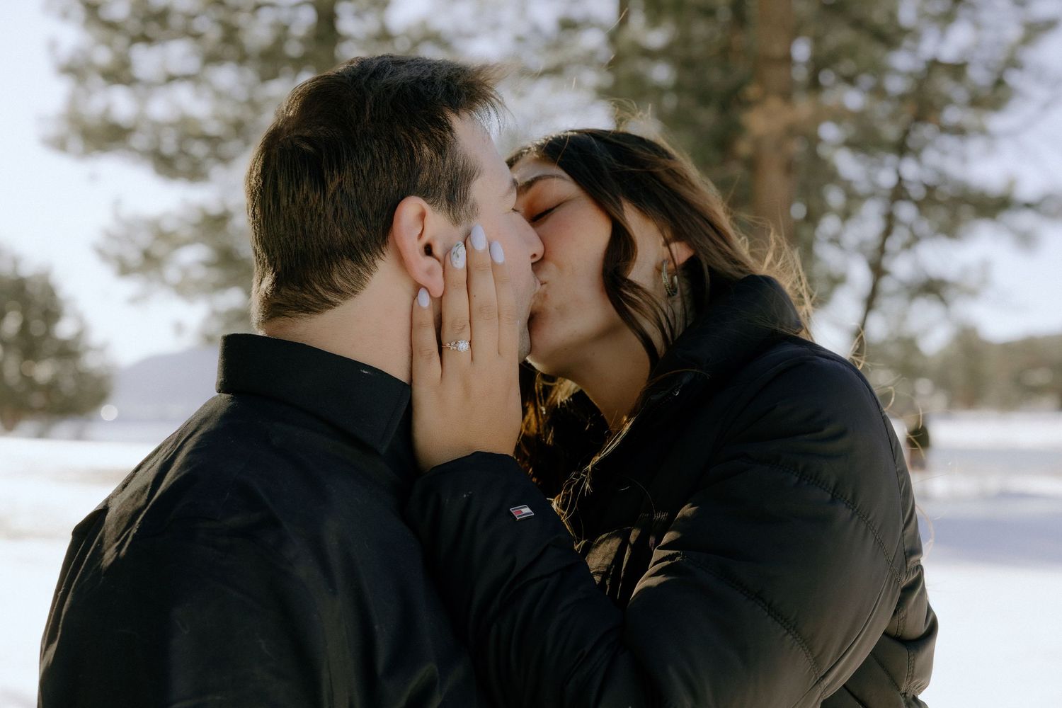 Couple sharing a romantic kiss outdoors in a snowy winter setting, woman displaying engagement ring.