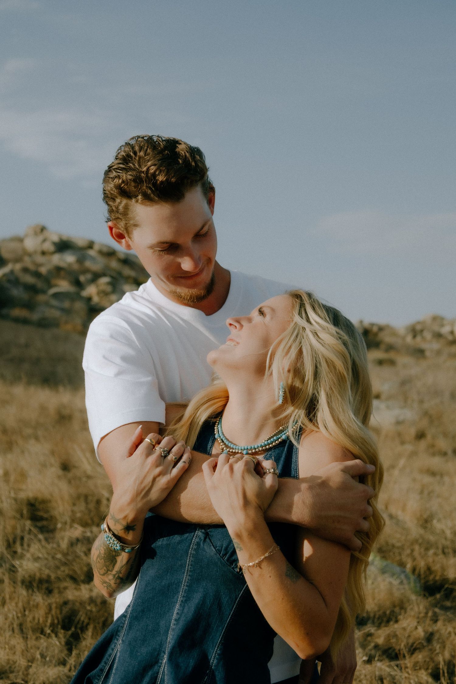 A couple embraces in a romantic moment against a desert landscape with rocky terrain and dry grass in the background.