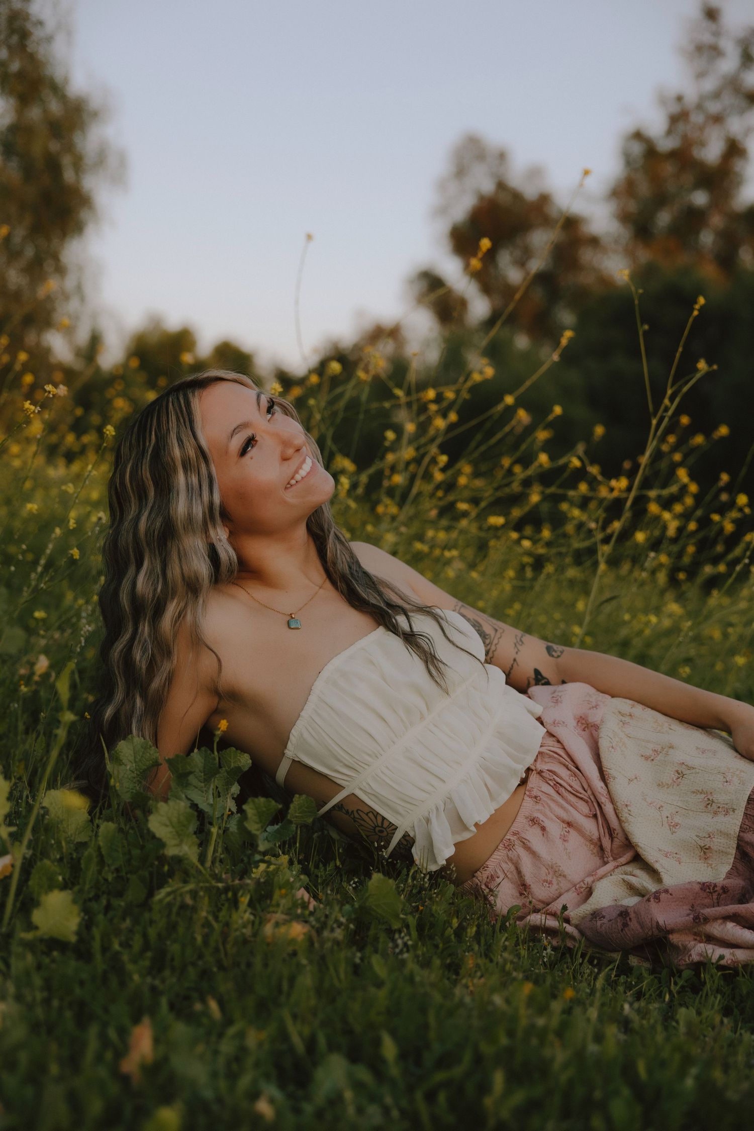 A person in a white top and pink skirt lies peacefully in a field of yellow wildflowers at golden hour.