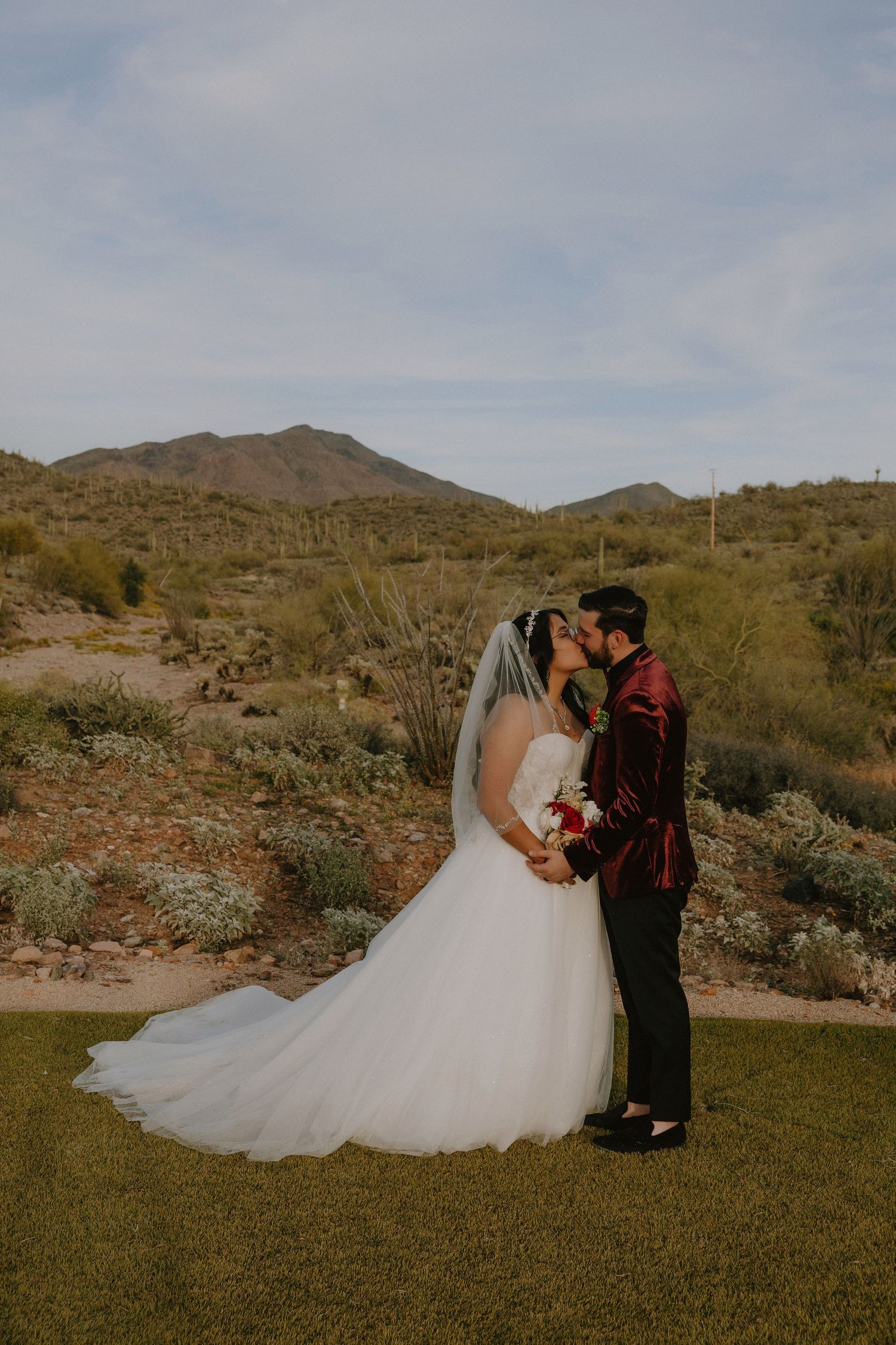 Bride and groom share a kiss in a white ballgown and burgundy suit with desert mountains backdrop.