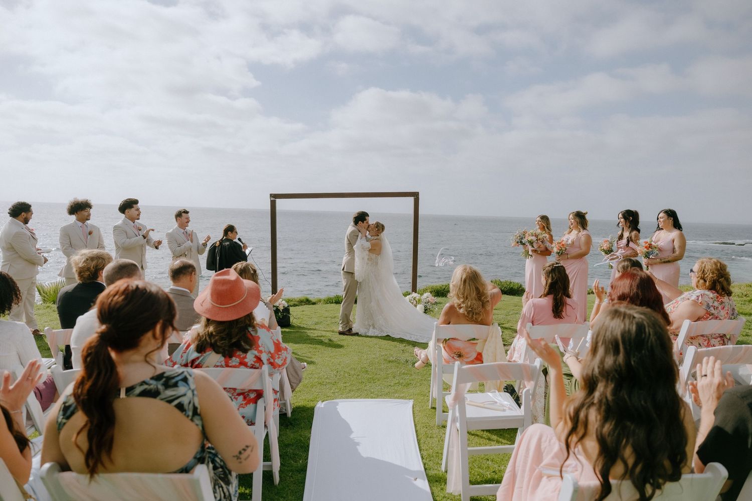 Outdoor oceanfront wedding ceremony with couple kissing at wooden arch, guests seated on white chairs on green lawn.