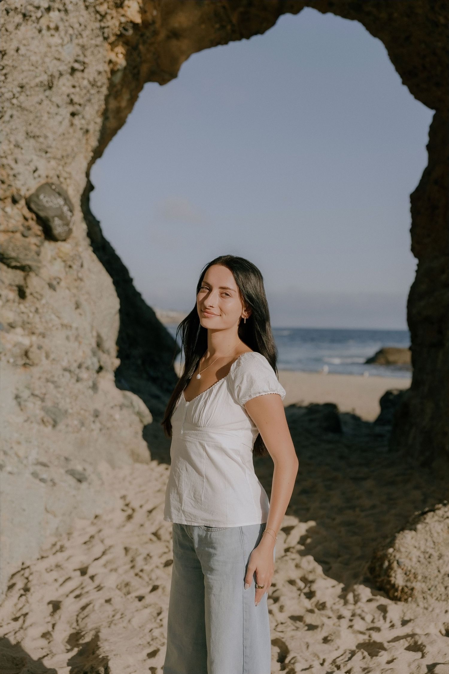A young woman in a white top and gray pants smiles near a rocky sea arch on a sandy beach.