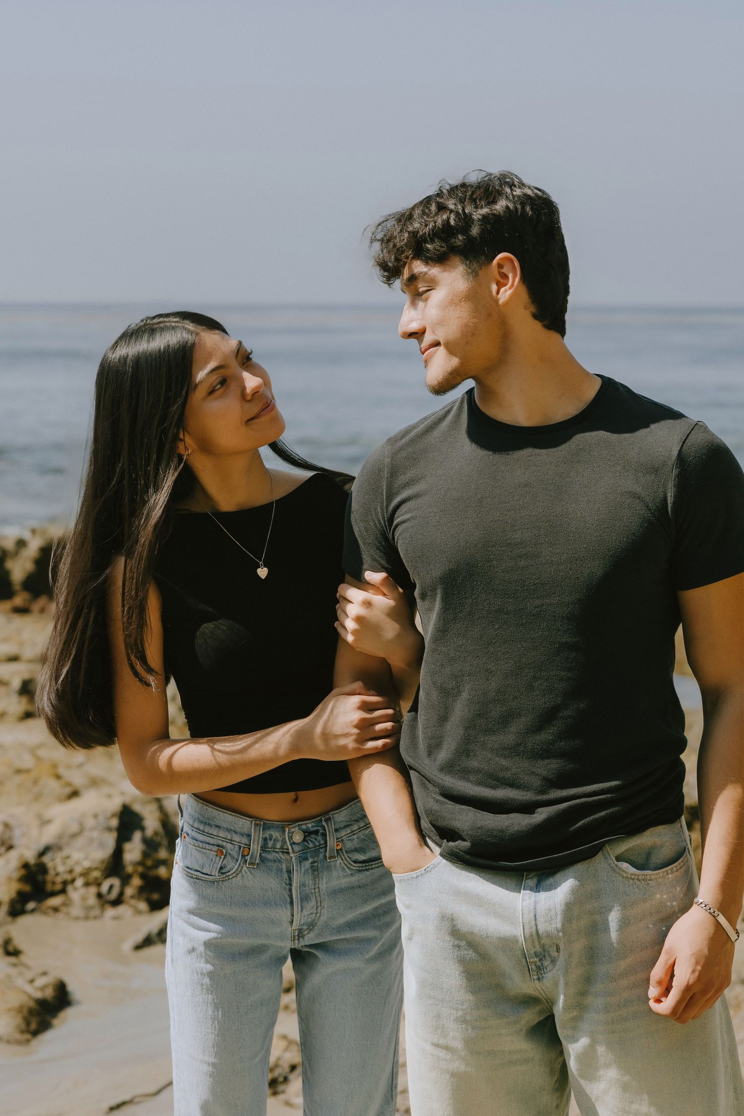 Young couple in matching black tops sharing a romantic moment on a rocky beach with ocean views.