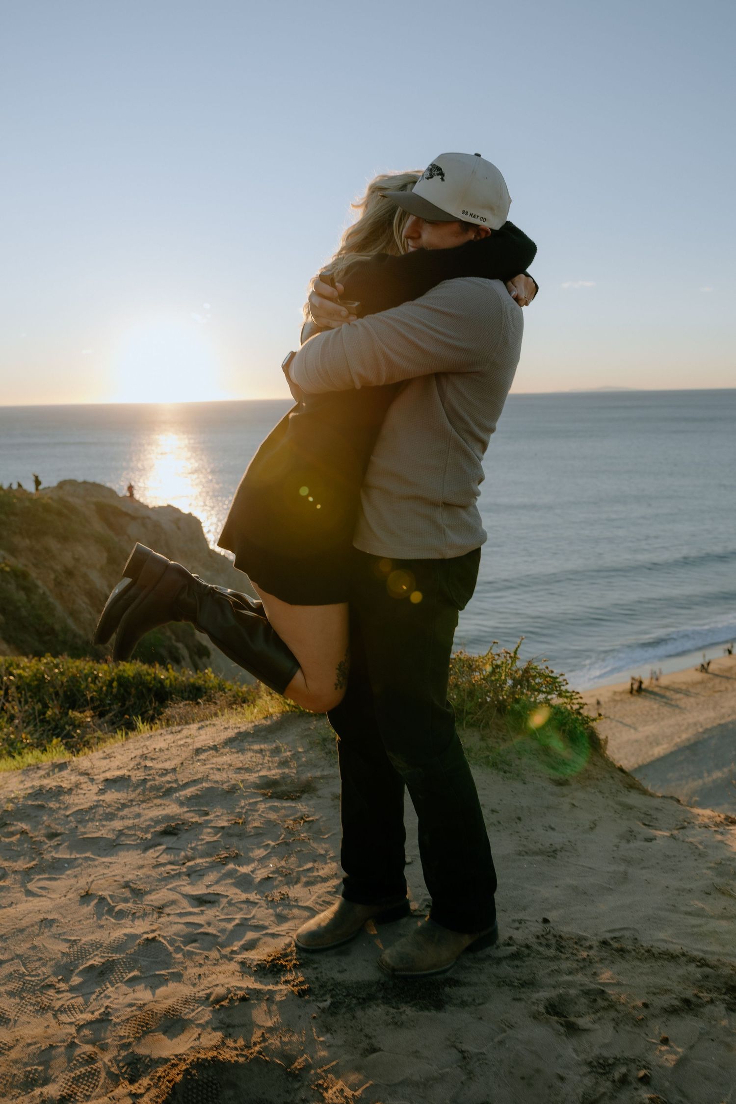 Couple embraces on coastal cliff during golden sunset over Pacific Ocean beach landscape.