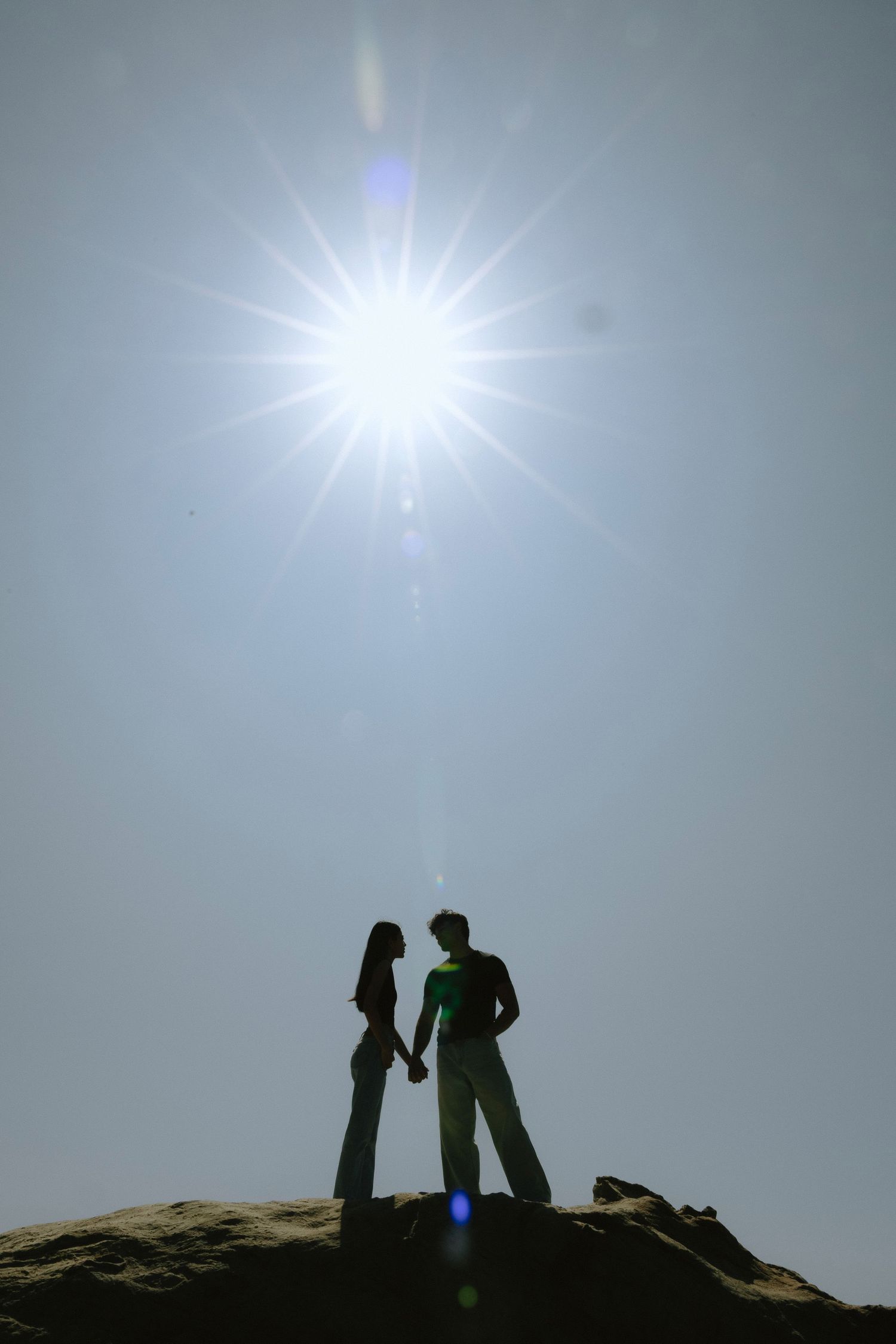 Silhouette of couple holding hands on rocky outcrop with bright sunburst in clear sky above them.