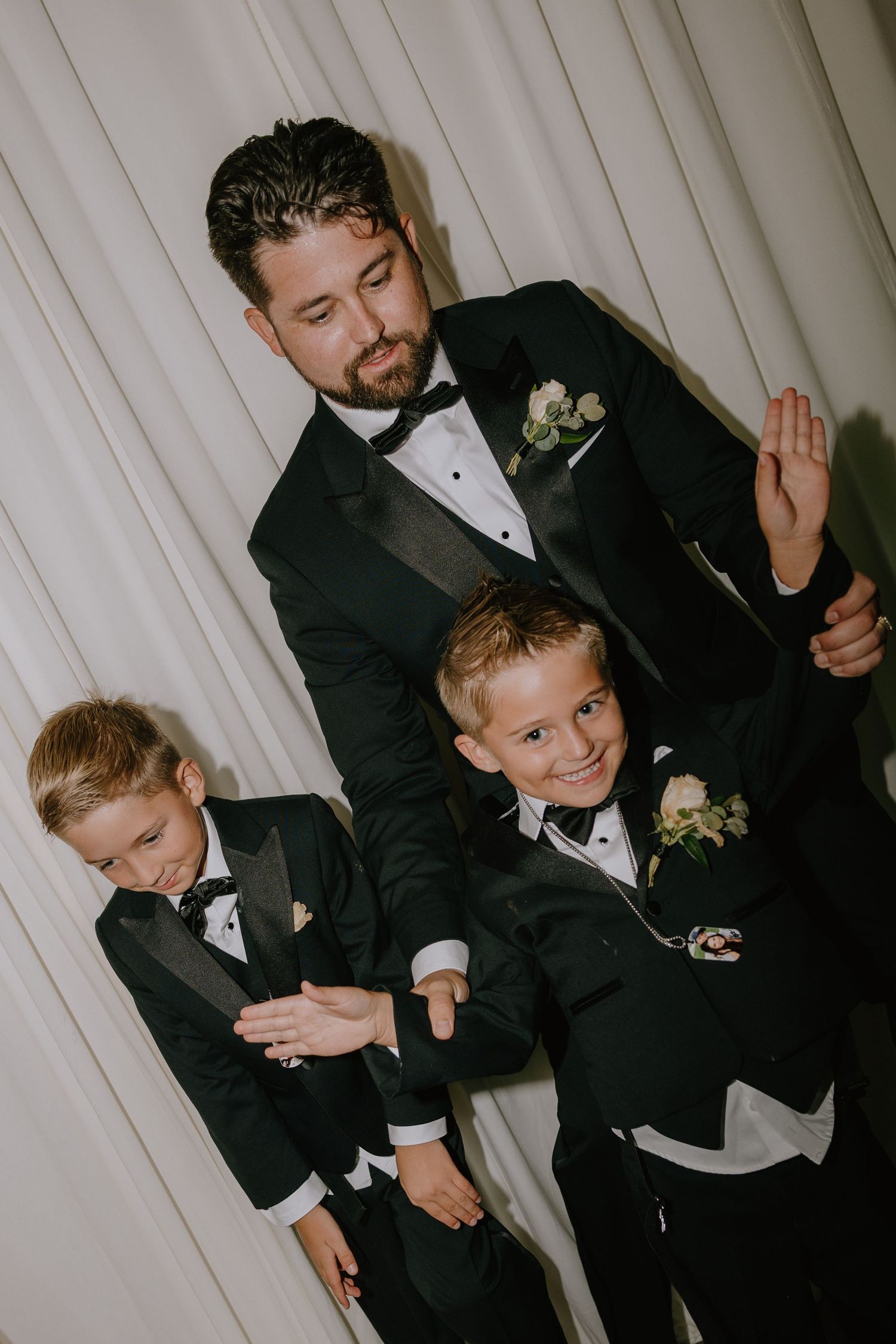 Groom in black tuxedo poses with two young boys in matching suits and boutonnieres at a wedding reception.