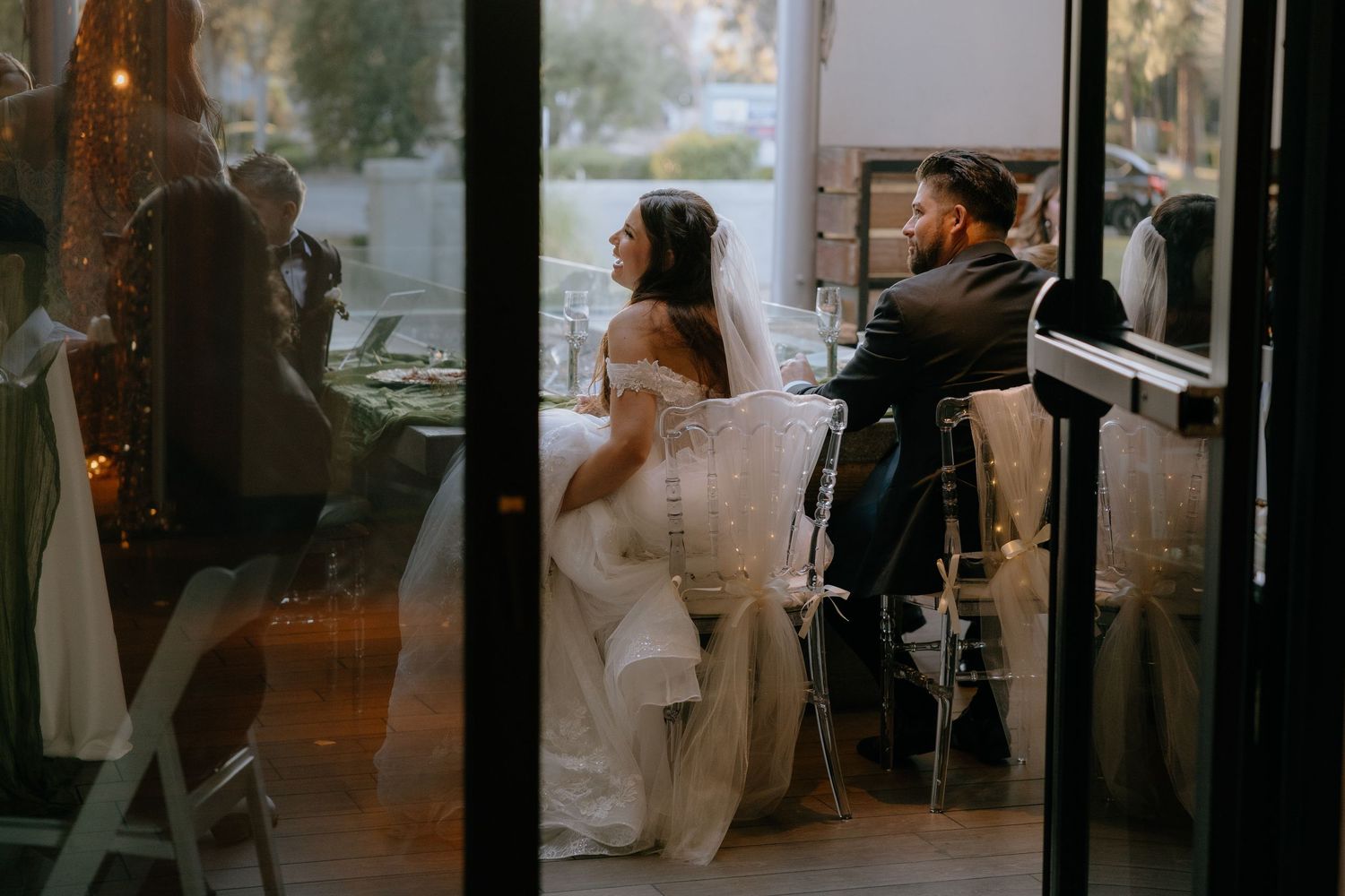 Bride and groom sit together at wedding reception table, viewed through glass doors, bride in elegant white gown.