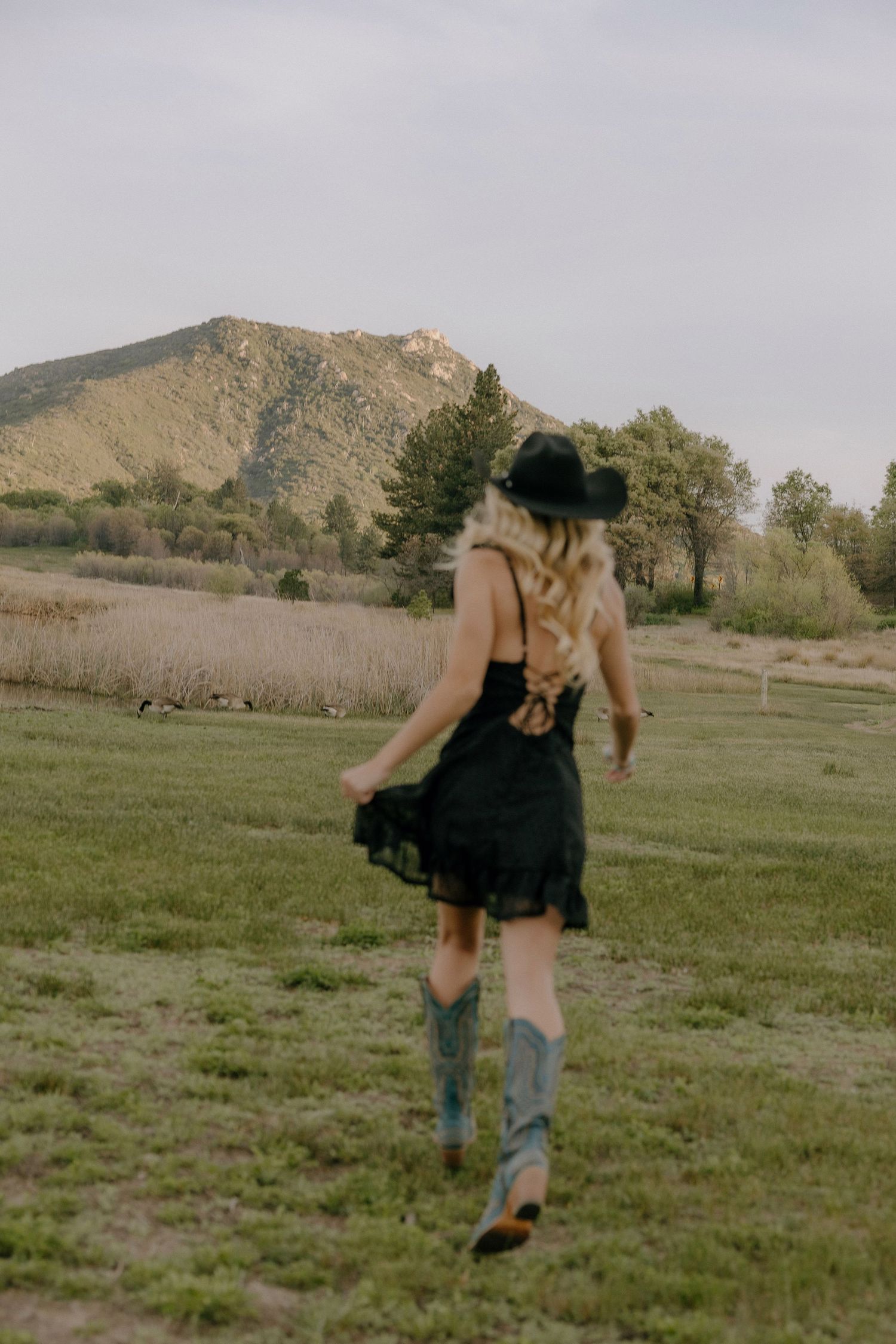 Blonde woman in black cowboy hat, lace dress, and turquoise boots walking through a green mountain meadow.