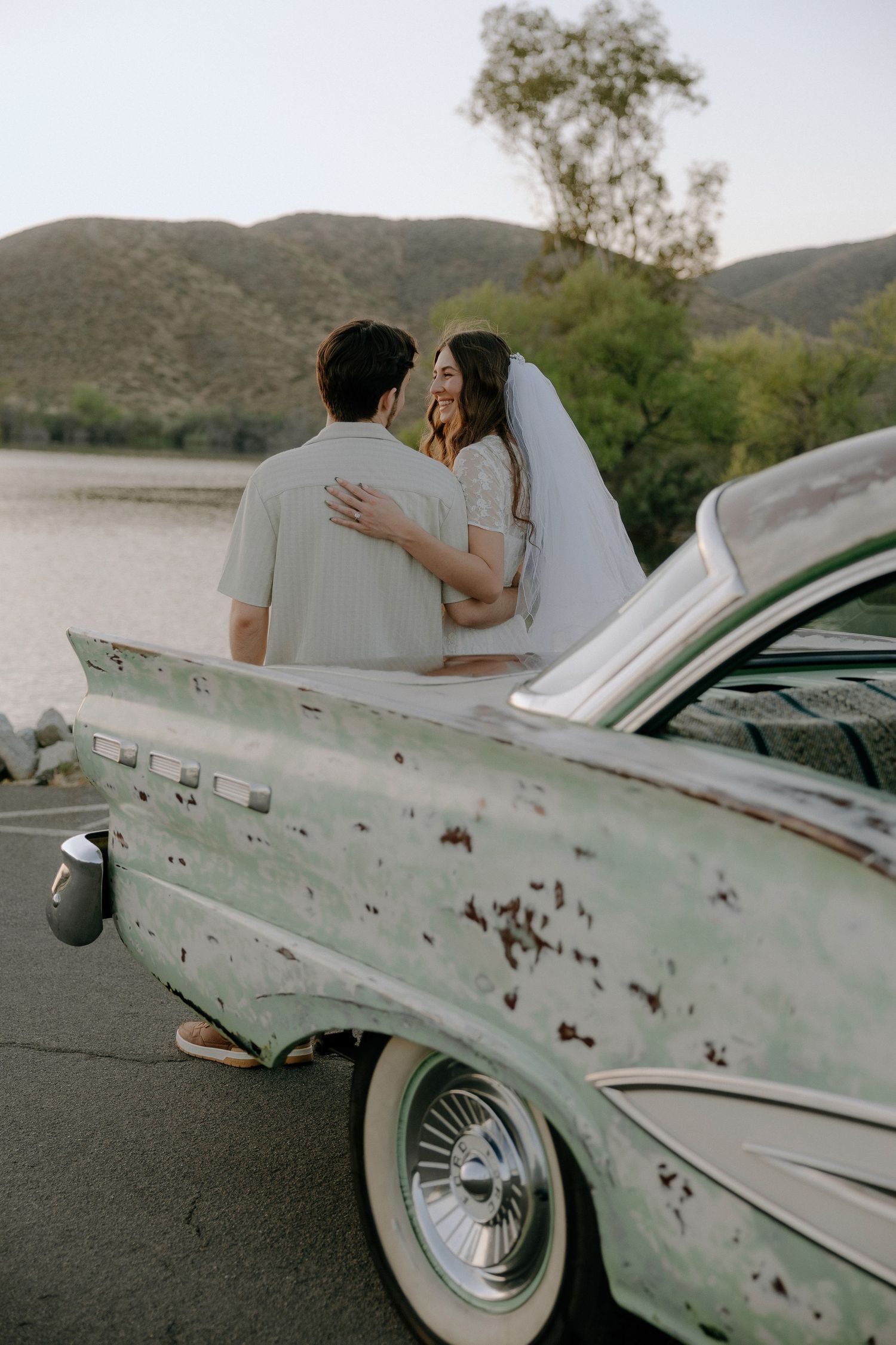 Bride and groom embracing beside a vintage mint green classic car with mountains and lake in background.