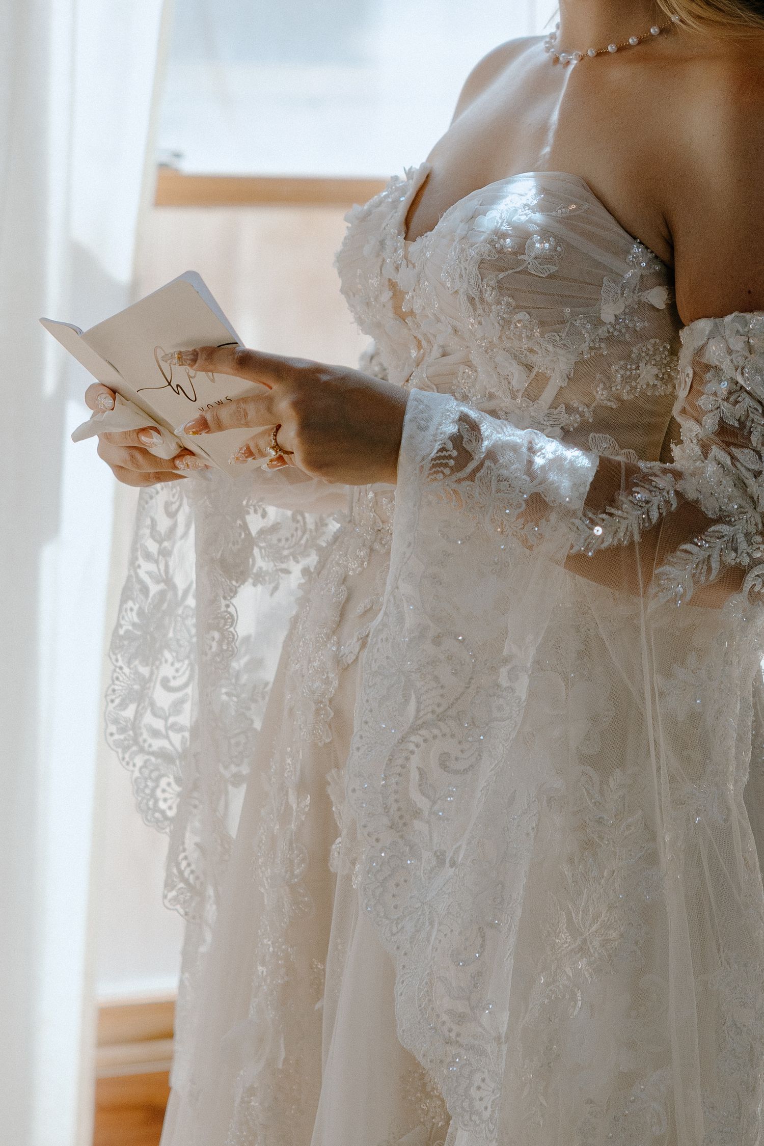 Bride in lace wedding gown reading a card by a window, wearing pearl necklace and elegant bridal attire.