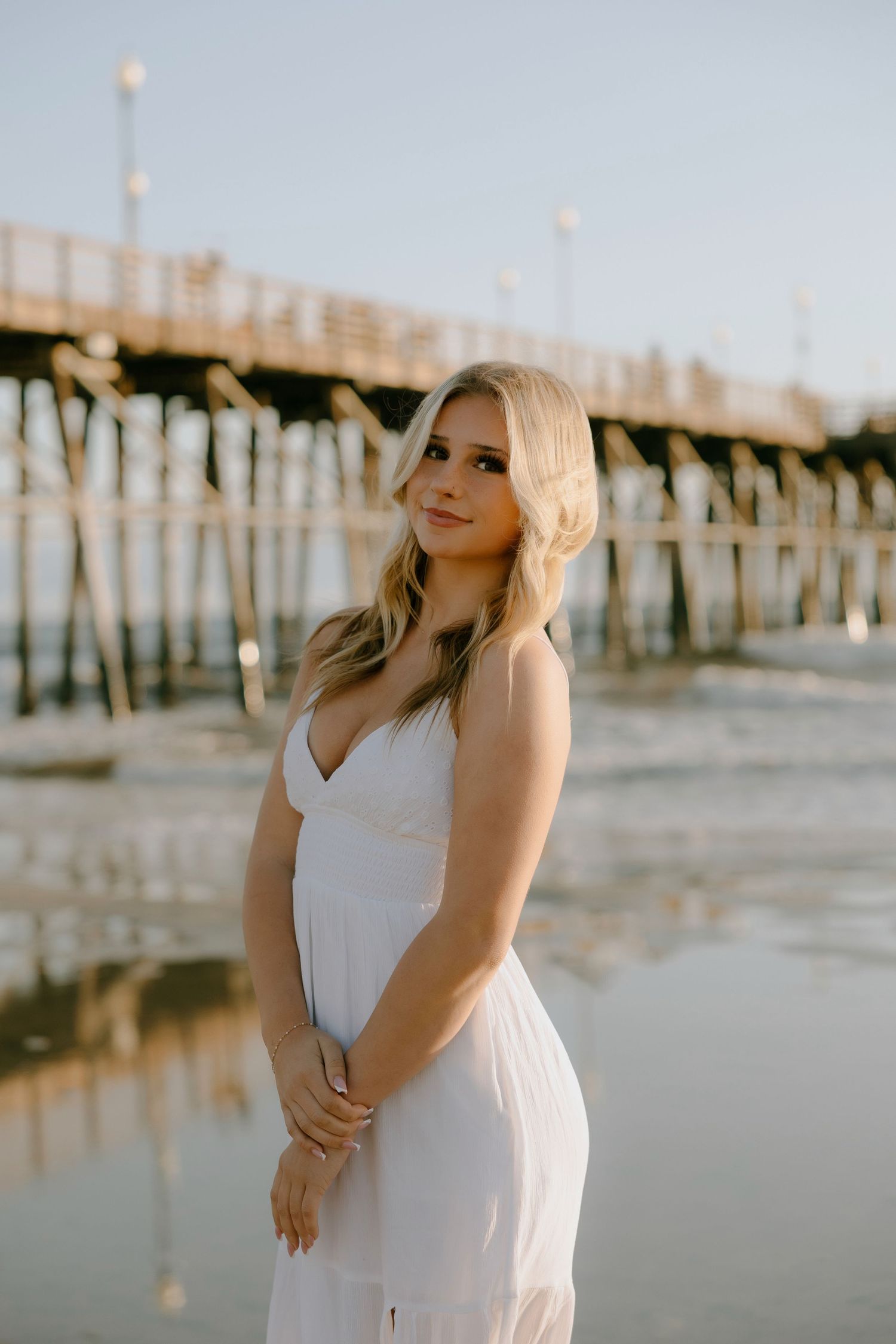 Blonde woman in white sundress posing near a wooden pier on a reflective beach at golden hour.