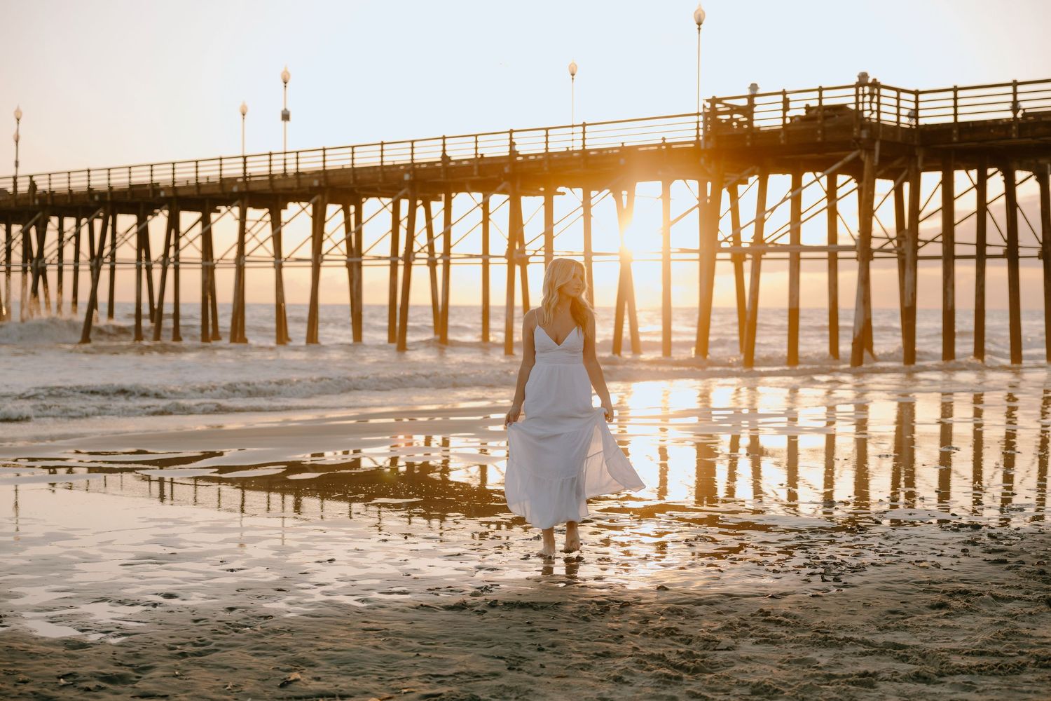 Woman in white dress walking on beach at golden sunset near a wooden pier with reflections on wet sand.