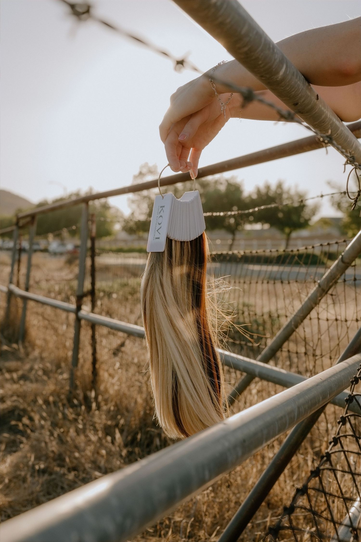 Hand holding a blonde hair extension swatch on a metal fence outdoors in a rustic setting.