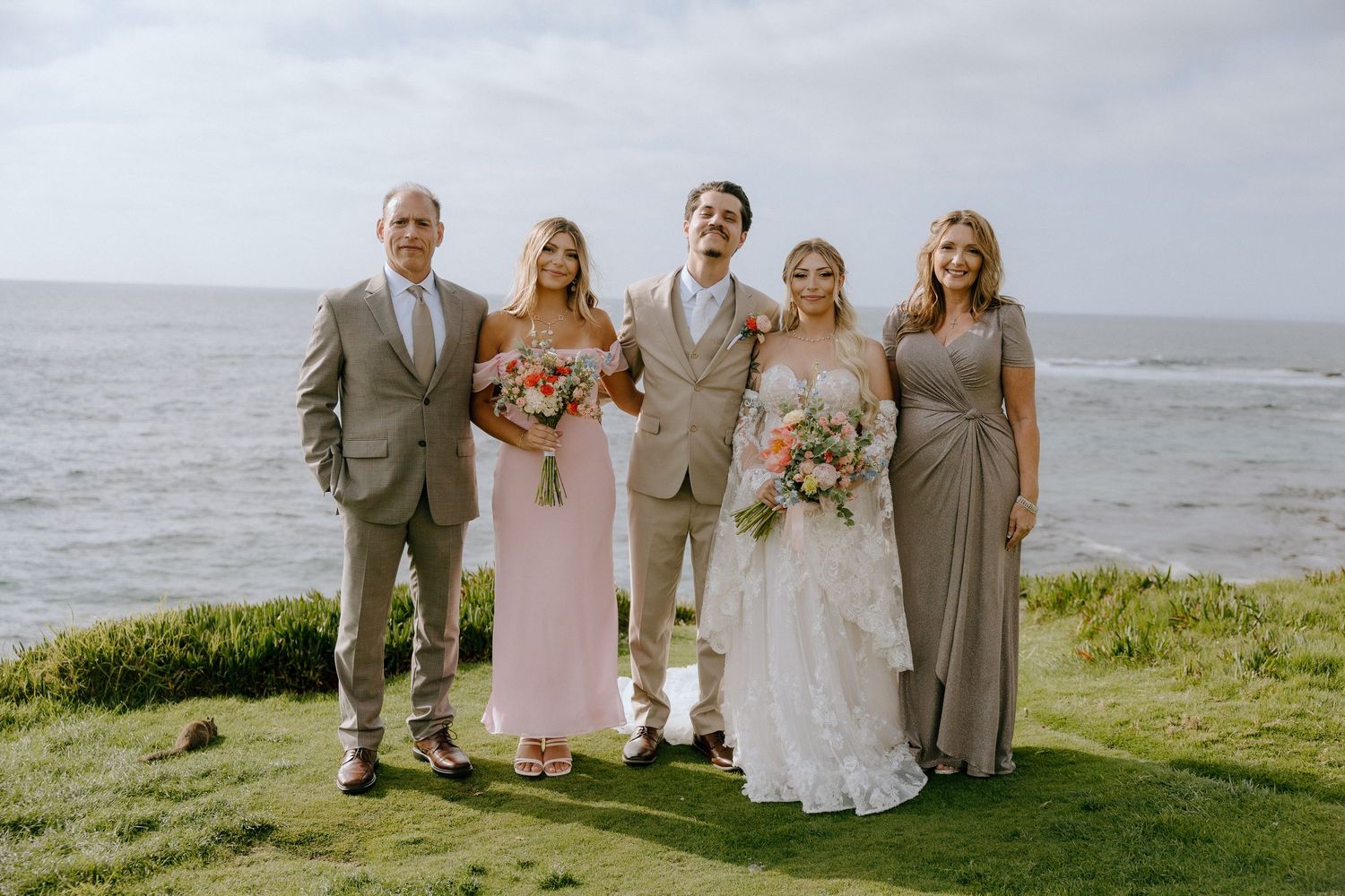 Wedding party poses oceanside, bride in white gown, bridesmaids in pink and taupe dresses, groomsmen in tan suits.